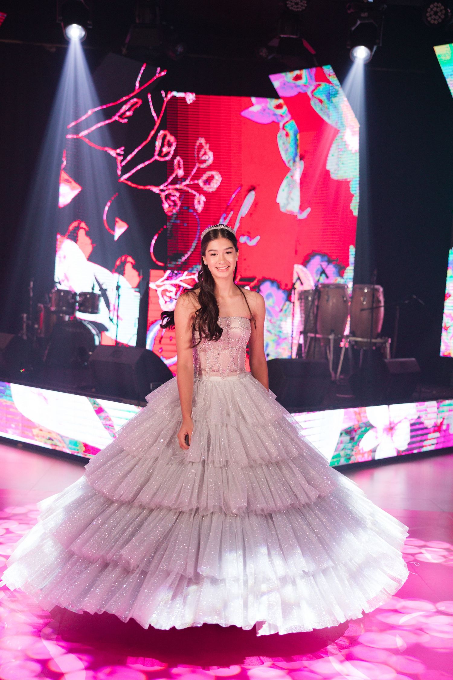 A person in a layered tulle ballgown poses on a pink-lit stage with dramatic lighting and video screens behind them.