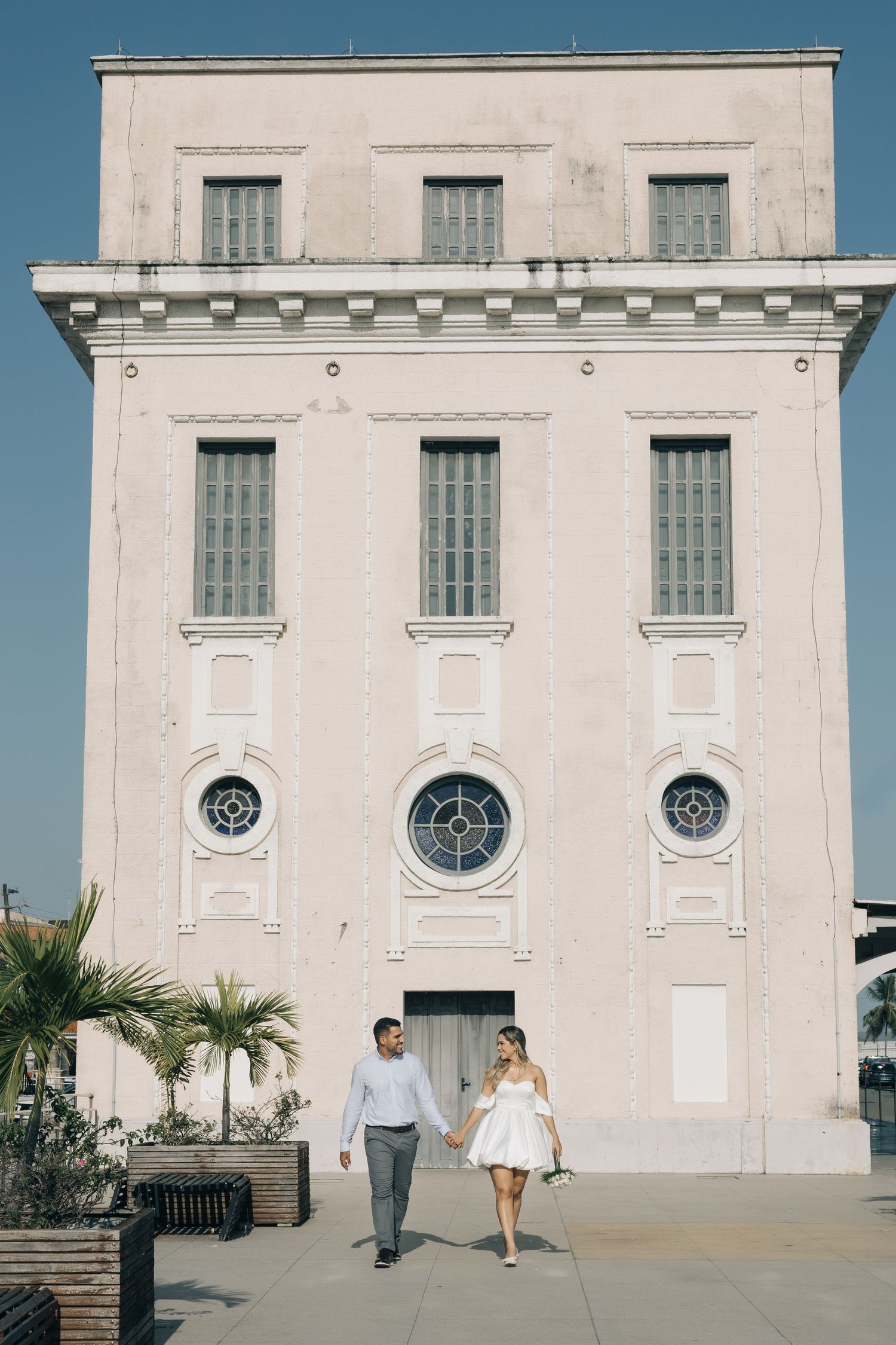 A couple walking hand in hand in front of a historic white building with palm trees.