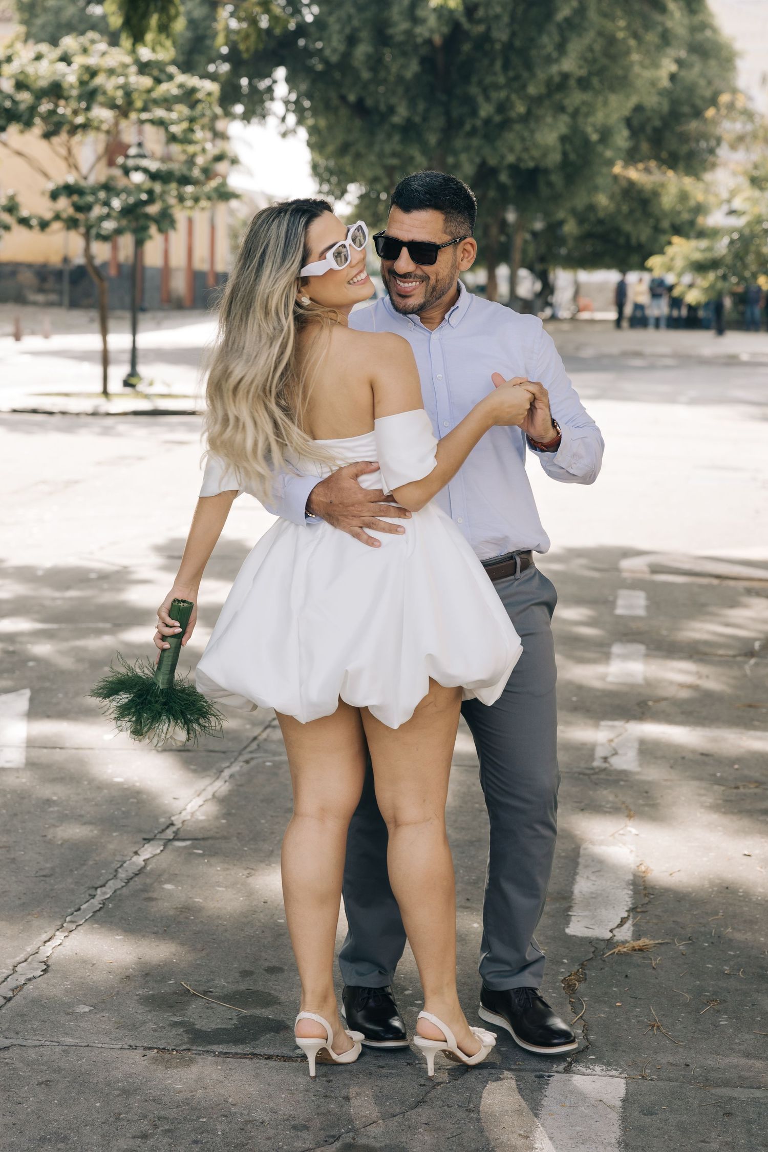 A couple sharing a romantic dance moment outdoors in casual attire, with sunlit trees creating dappled shadows.
