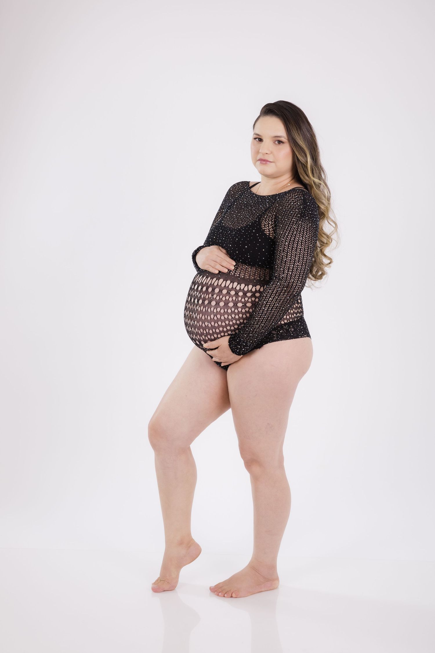 A pregnant woman poses in sheer lace bodysuit during a maternity photoshoot against a white studio background.