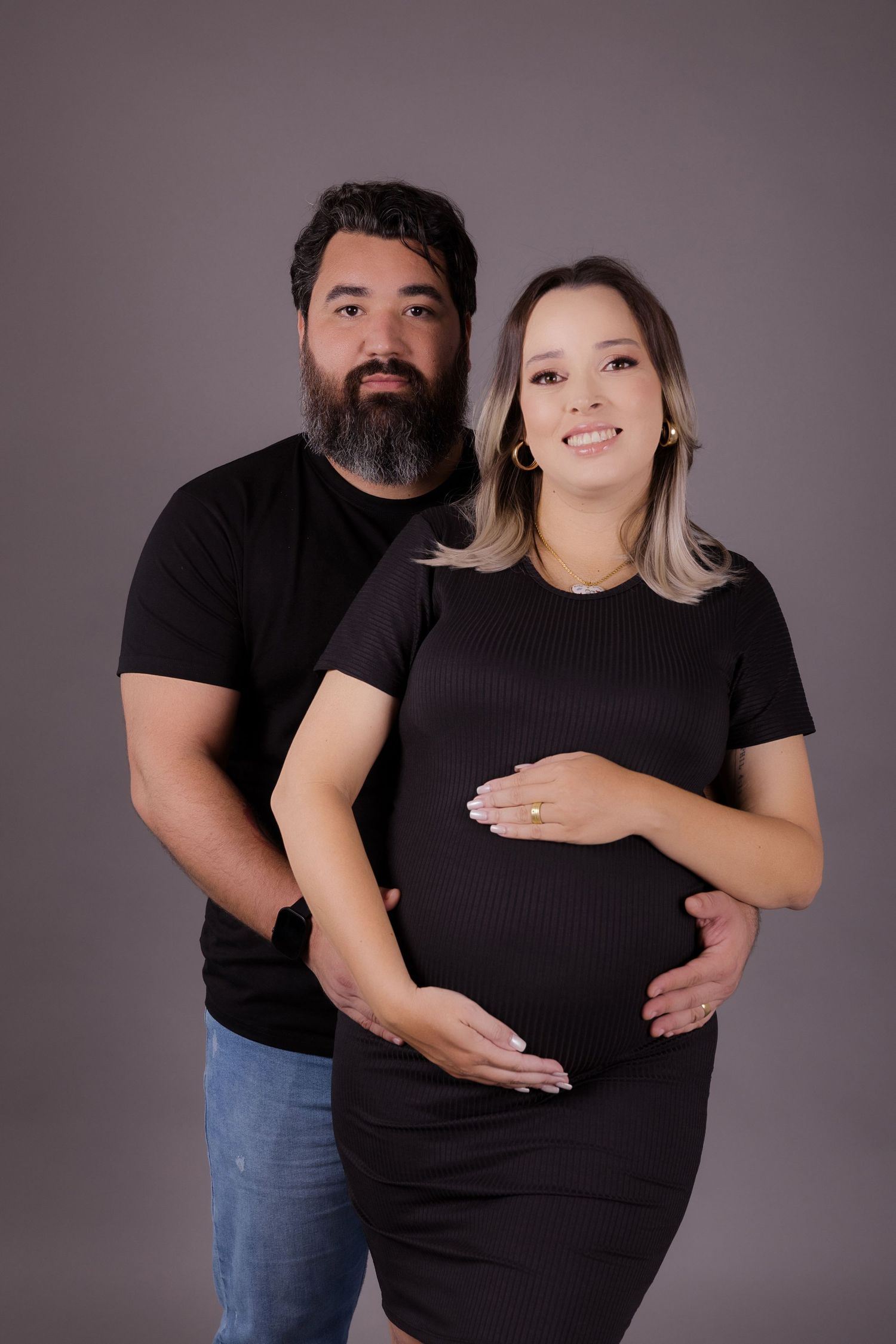 A couple wearing black shirts poses for a pregnancy portrait against a dark grey studio background.