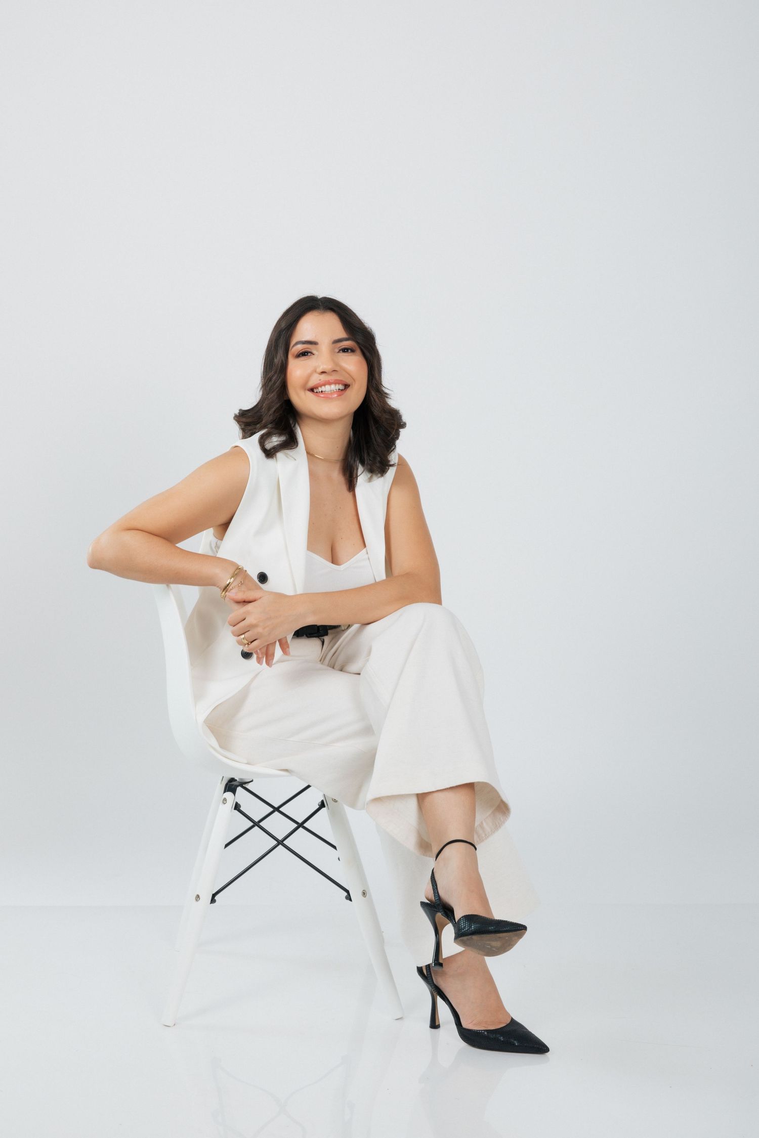 Model in white outfit and black heels poses elegantly on white stool against light background.