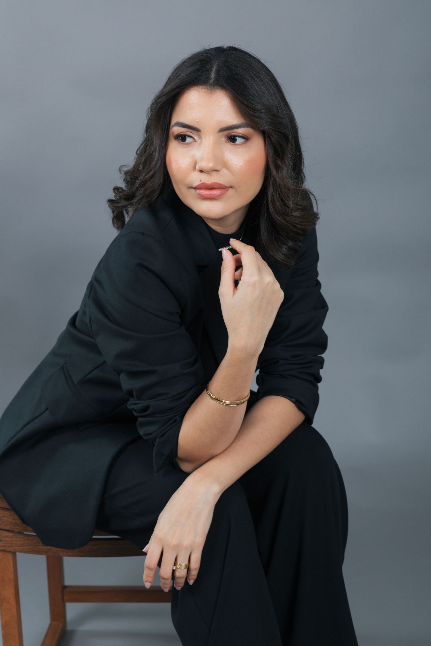 Professional studio portrait of someone sitting elegantly in black business attire against a gray backdrop.