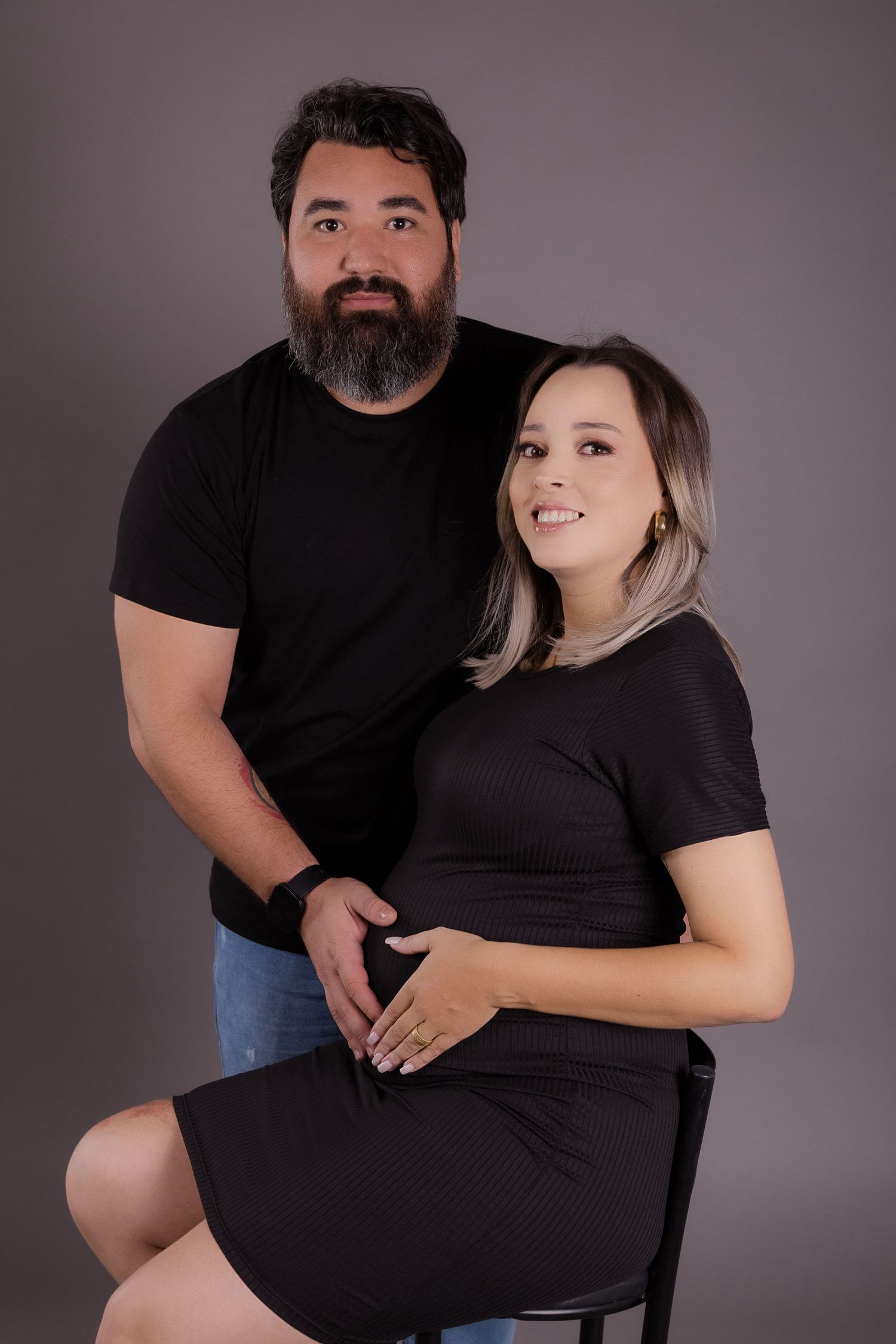 Expecting couple in dark clothing pose together against a gray studio backdrop.