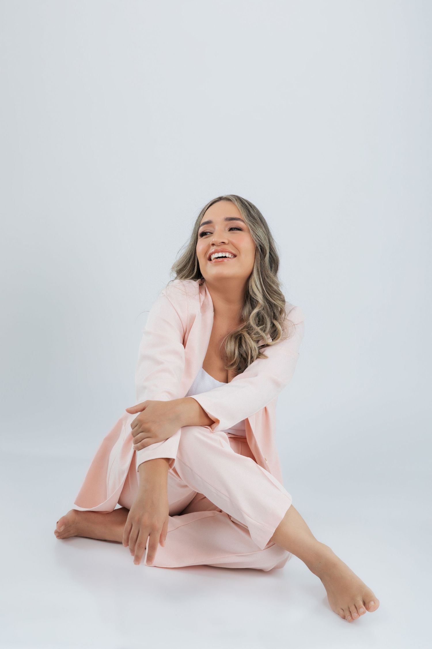 A person wearing a pink pantsuit sits casually on a white floor with a joyful expression against a light background.
