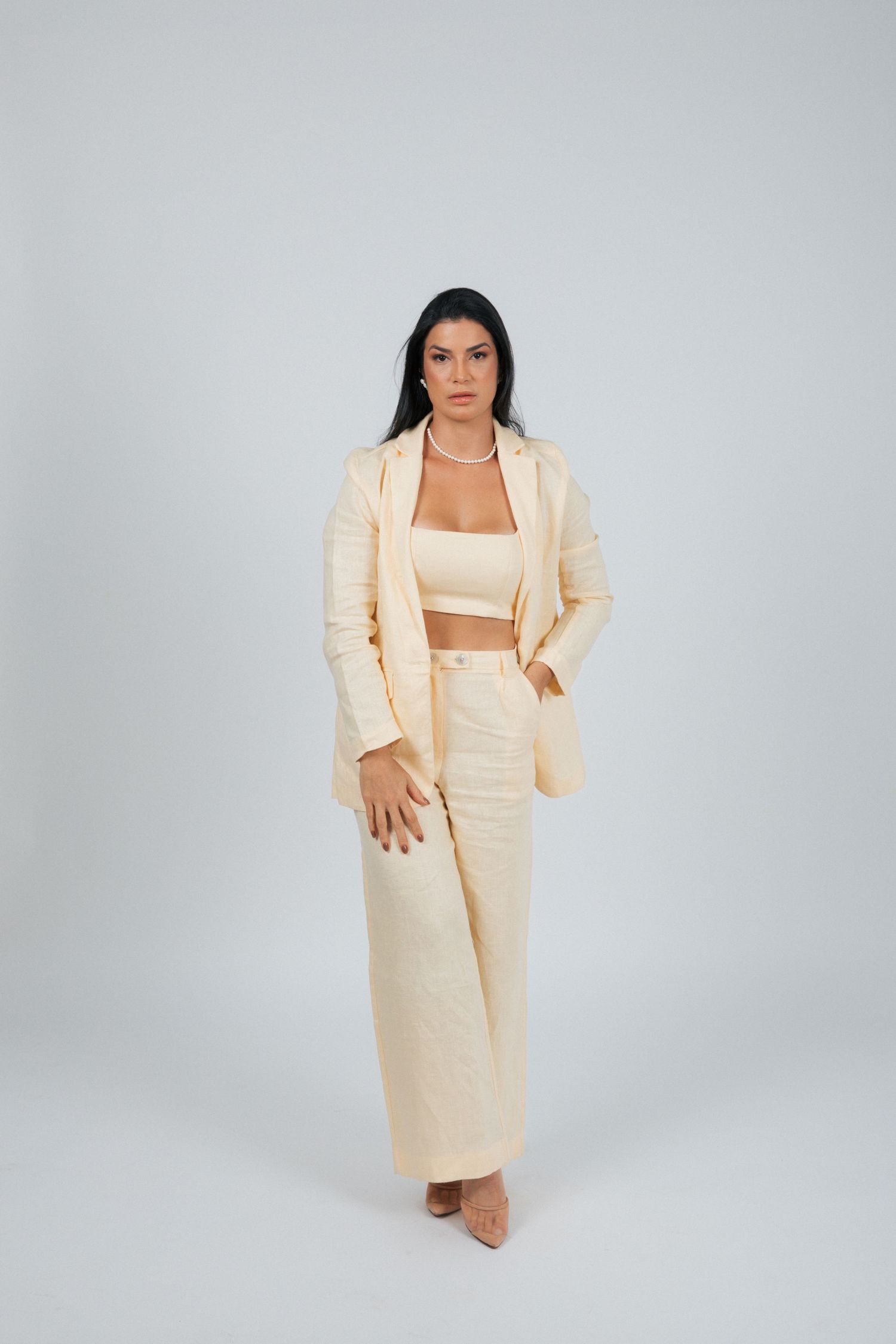 Person in light colored business suit and crop top stands confidently against grey backdrop.