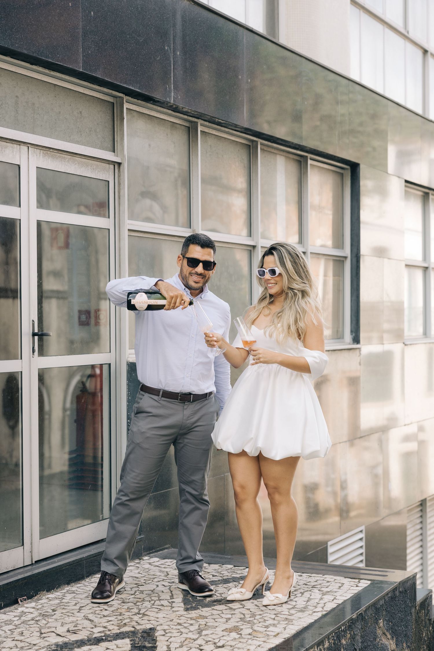 A couple pours champagne while standing on a patterned stone ledge outside a modern building.