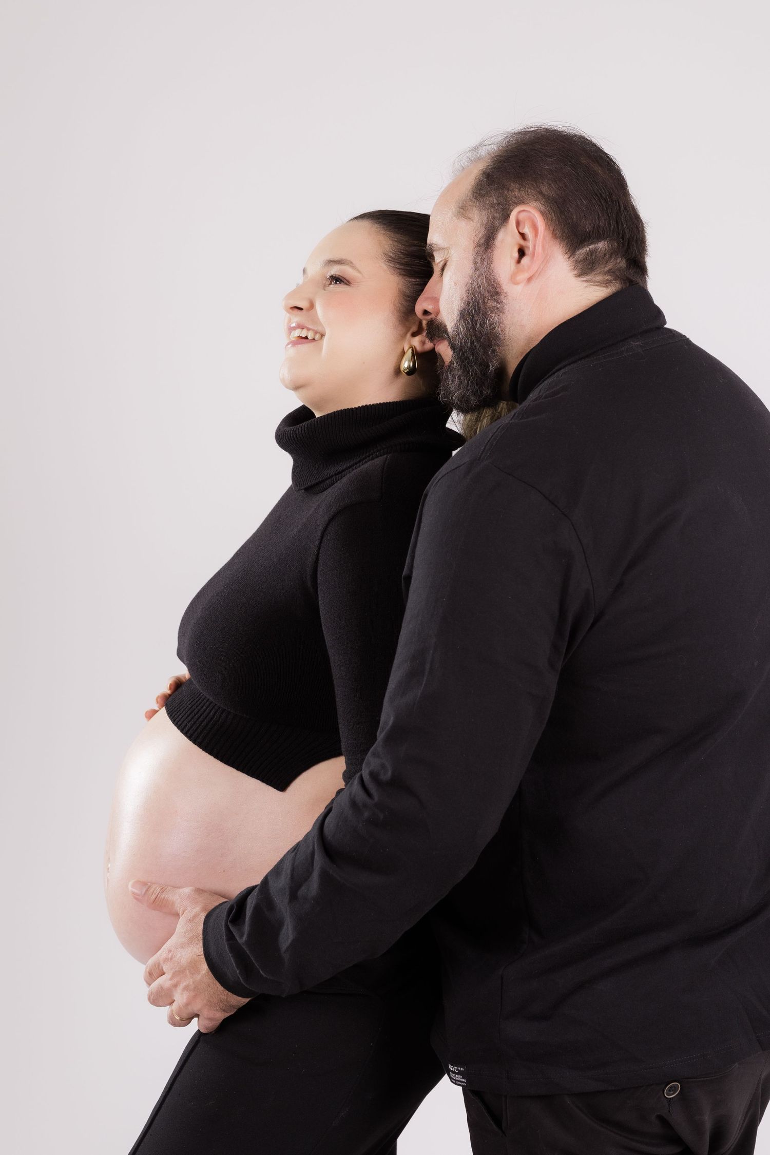 Intimate maternity photo of couple in black clothing embracing against white backdrop as they anticipate their new arrival.