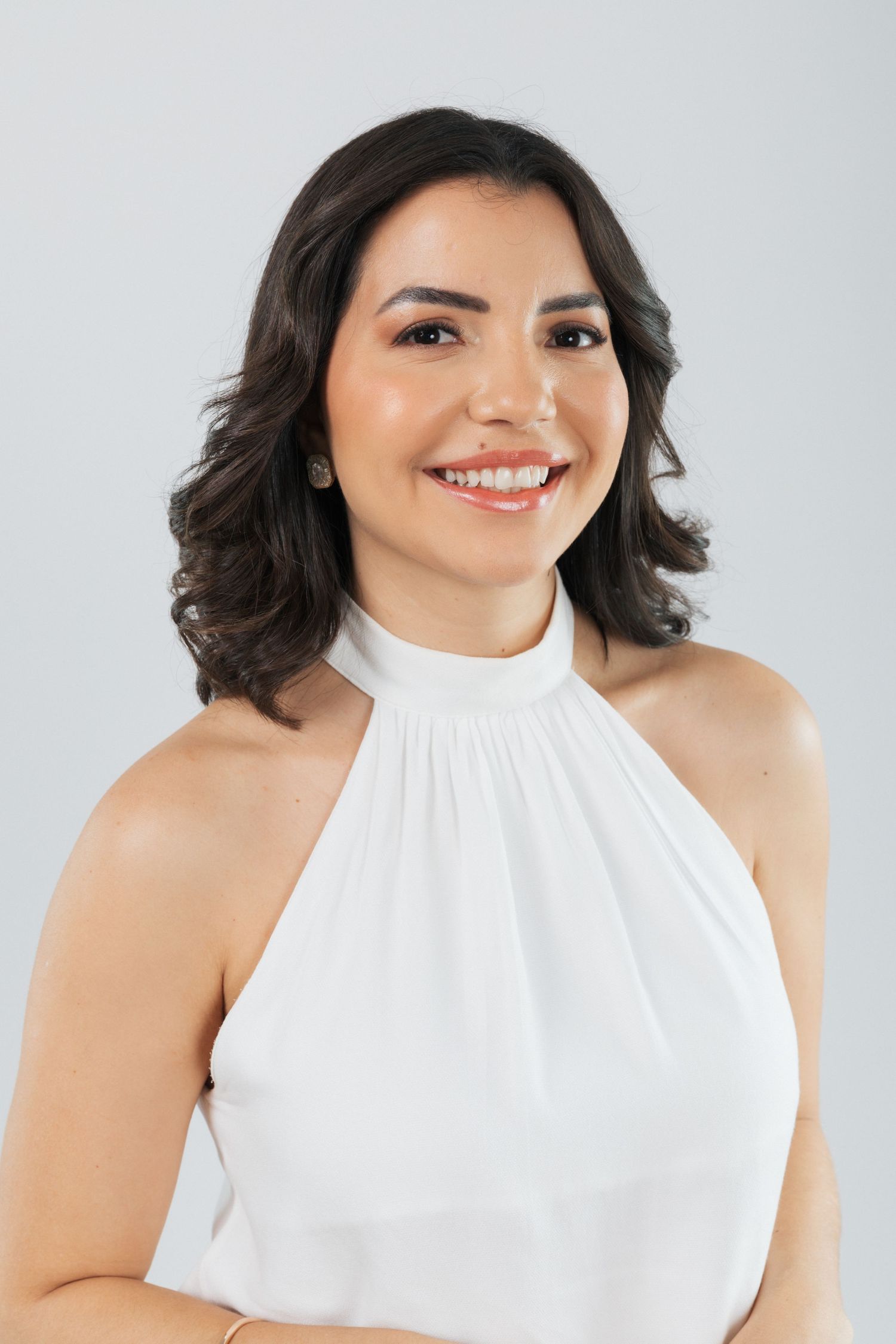 Professional portrait featuring a white halter top and dark wavy hair against a light gray background.