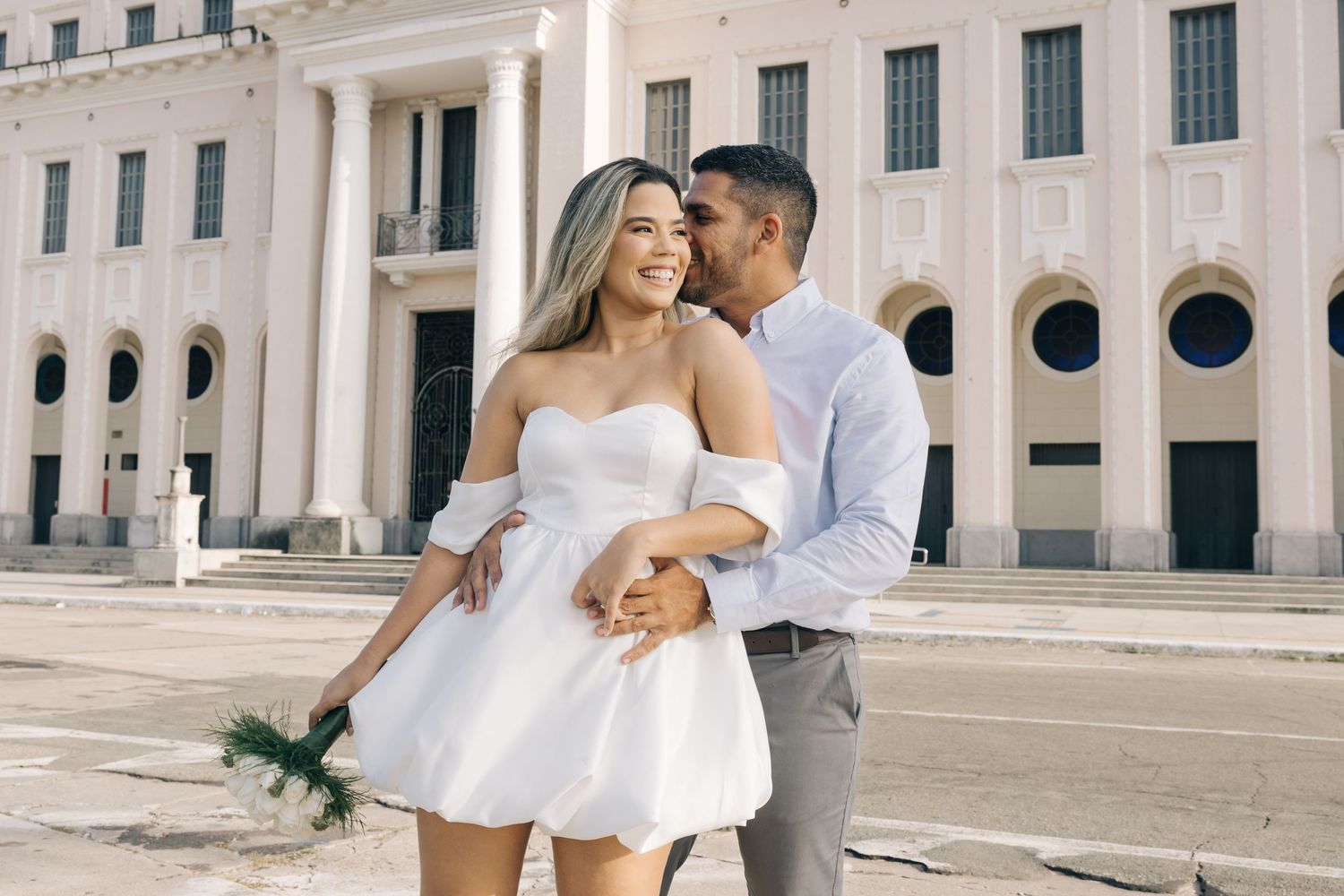 A romantic couple in wedding attire embrace and laugh together outside a grand stone building with columns.