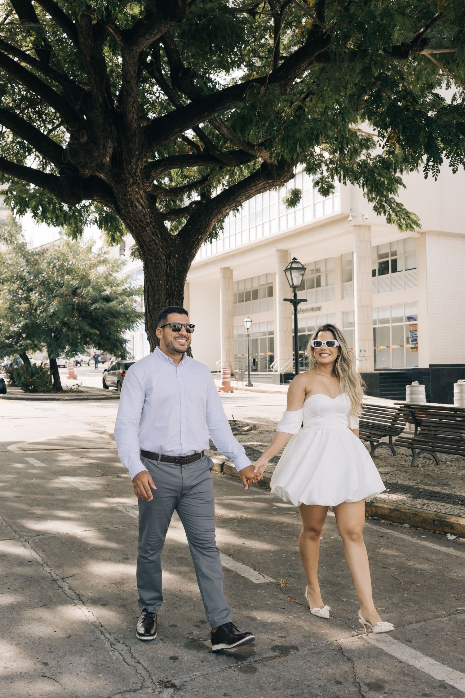 A couple walking hand in hand along a tree-shaded path in front of a white building.