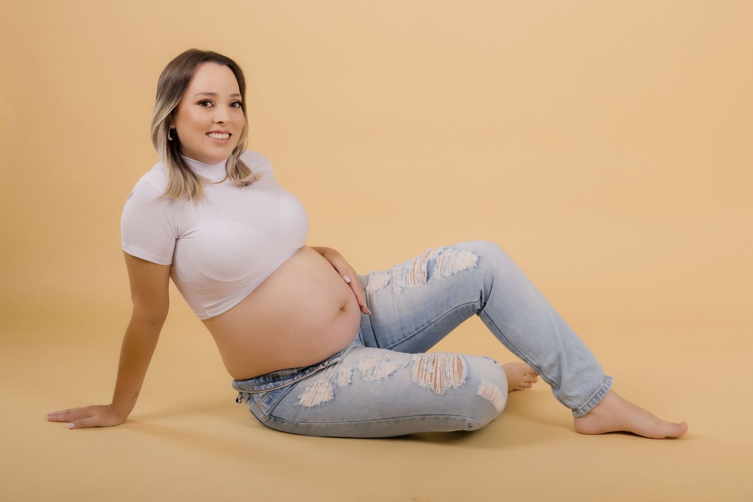 A pregnant woman in a white crop top and distressed jeans poses on a beige backdrop during a maternity photoshoot.