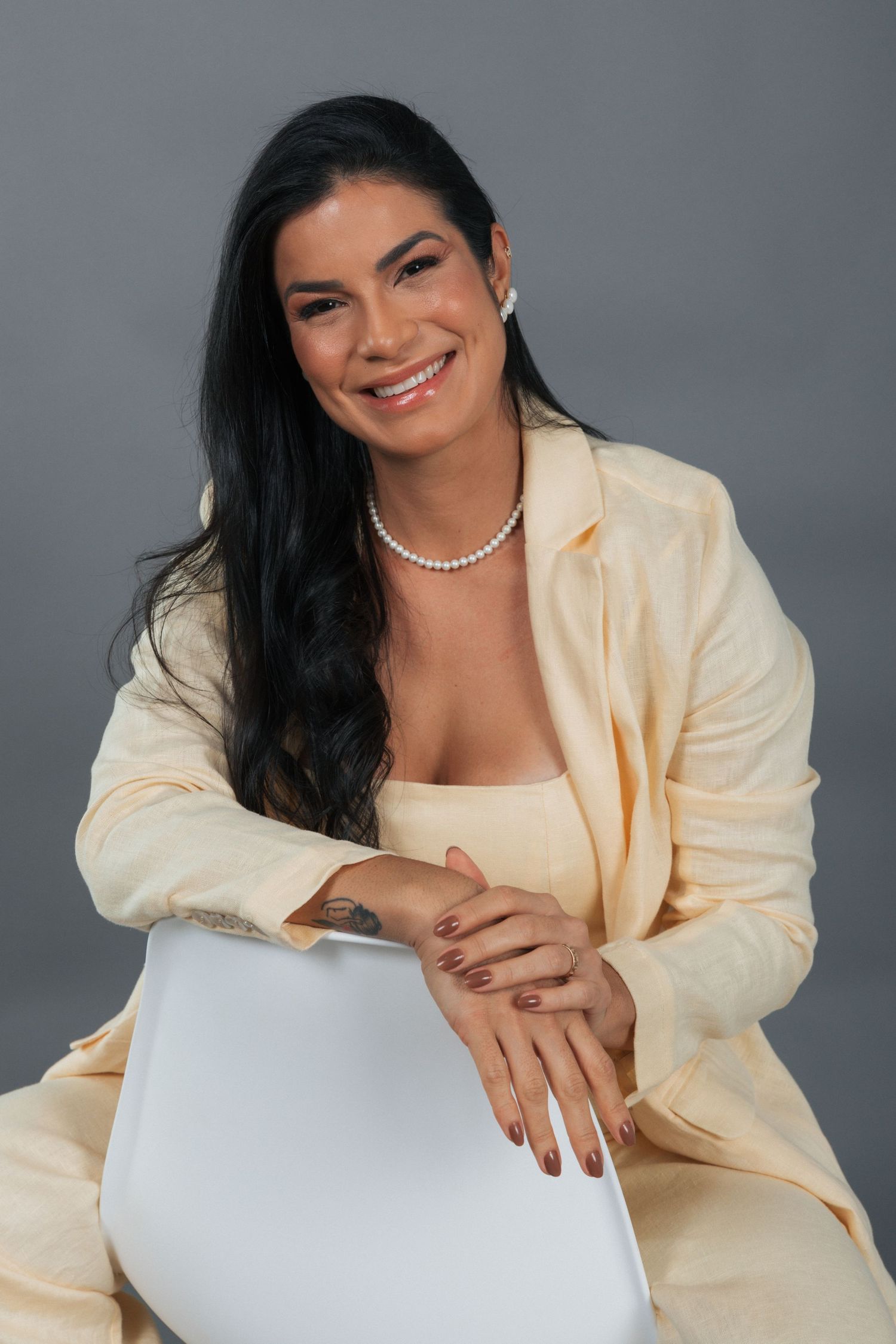 Professional studio portrait of a person with long dark hair wearing a cream blazer and pearl necklace against gray background.