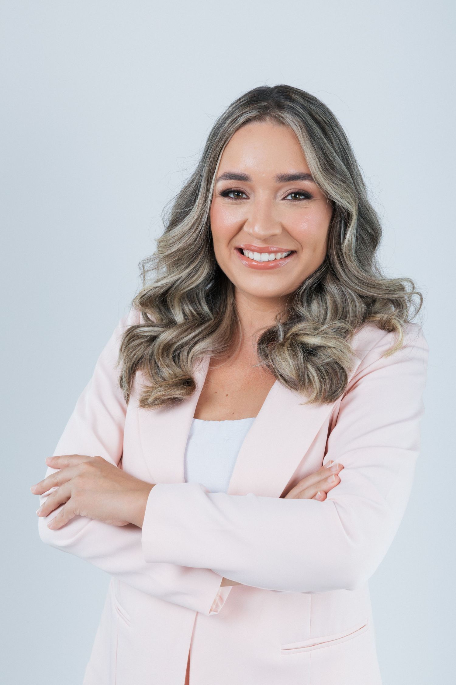 Professional woman in pink blazer with wavy silver-gray hair standing with crossed arms against light background.
