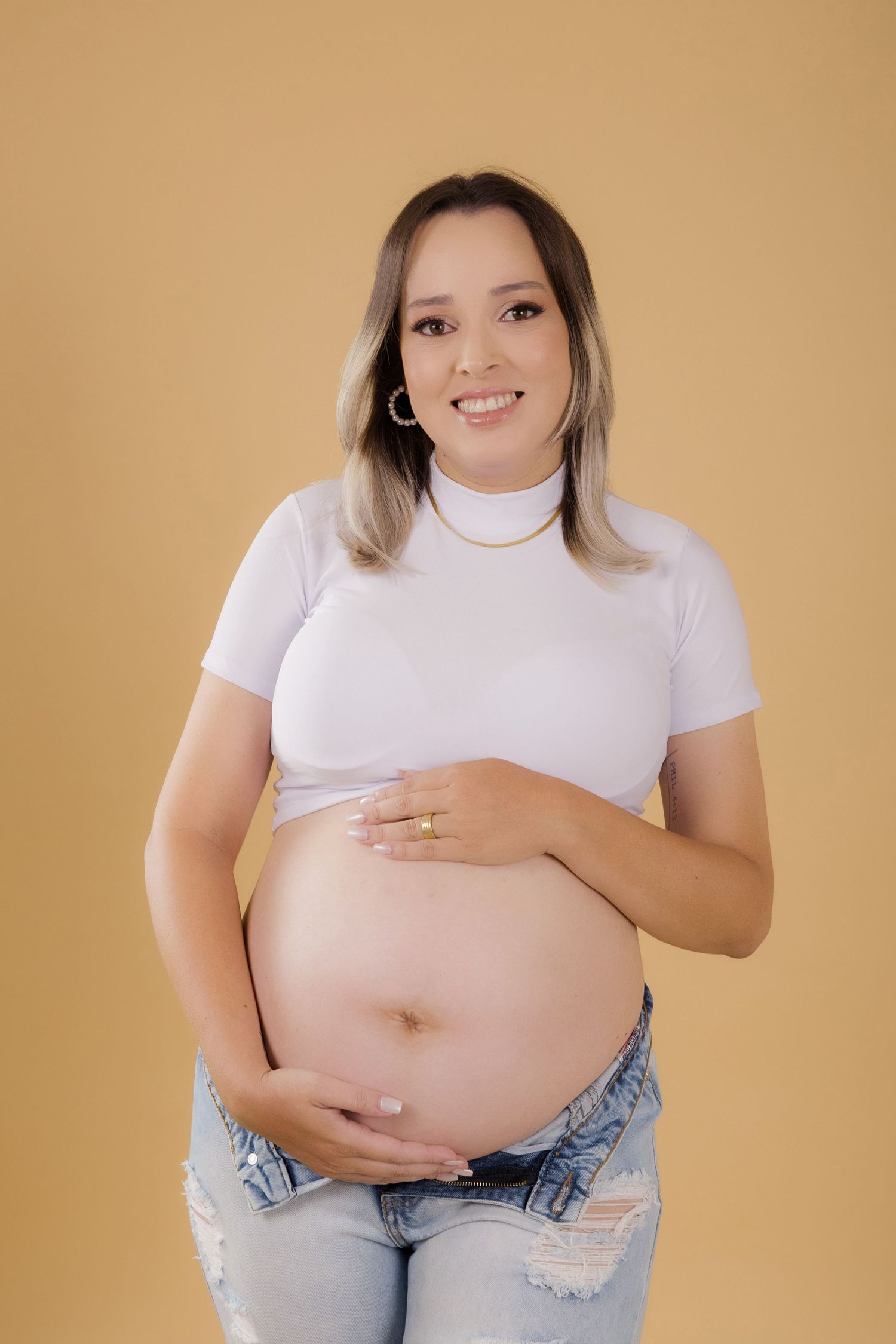 A pregnant woman in a white turtleneck and distressed jeans poses against a beige backdrop holding her belly.