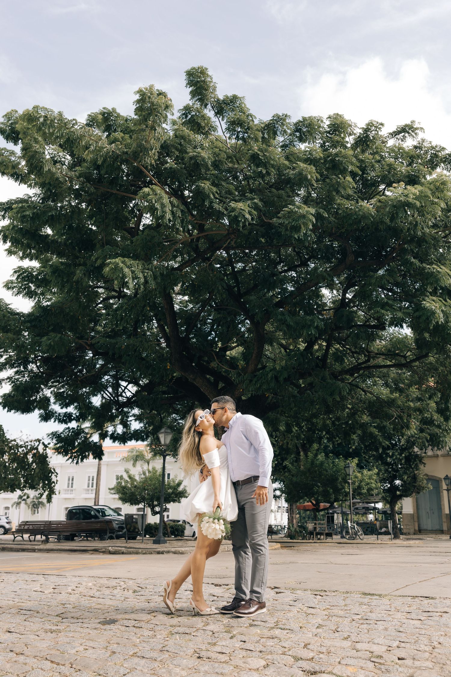 A couple shares a romantic moment on a cobblestone street underneath a large leafy tree on a cloudy day.