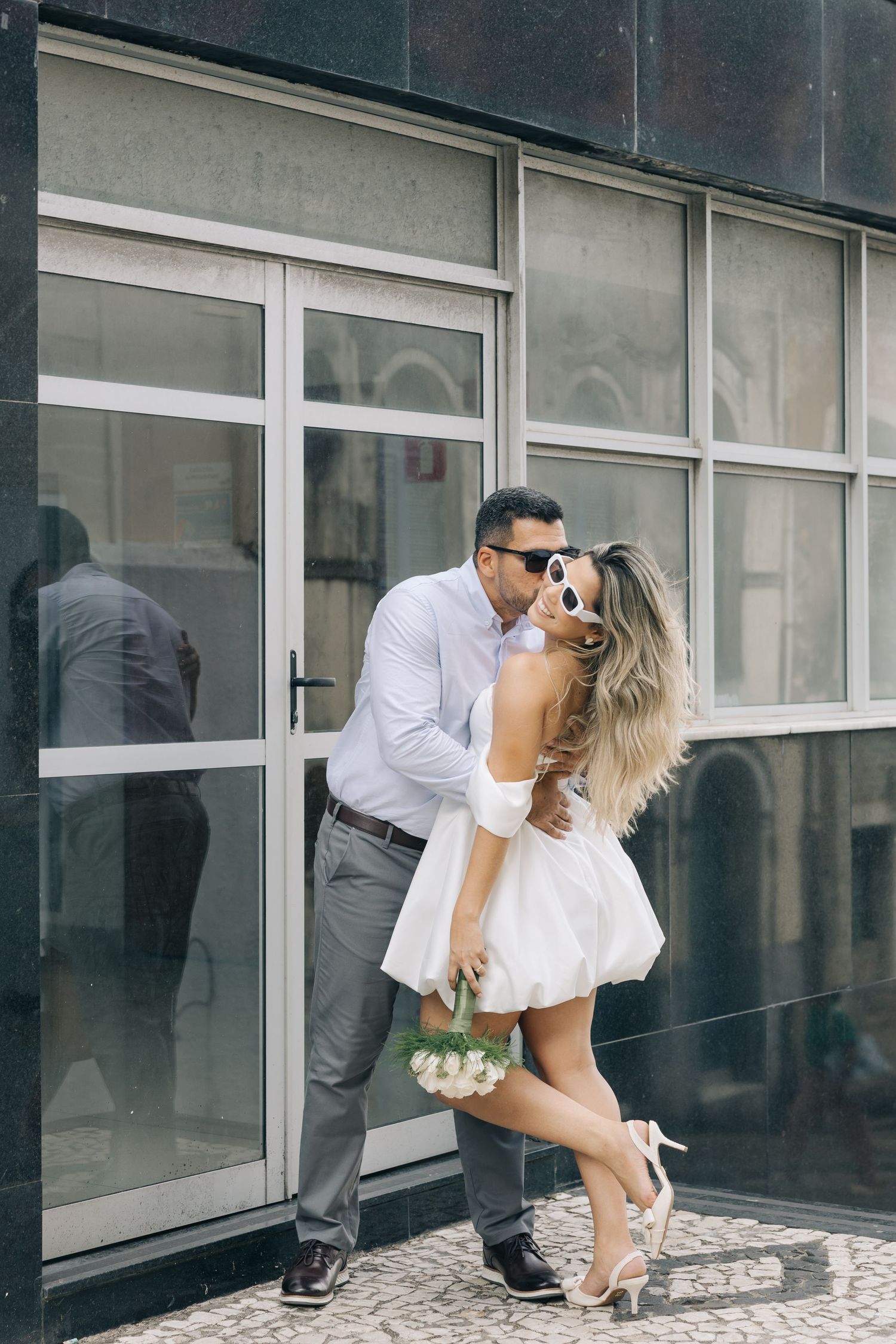 A romantic couple poses playfully outside a modern glass building, with her in a short white dress and him in business casual.