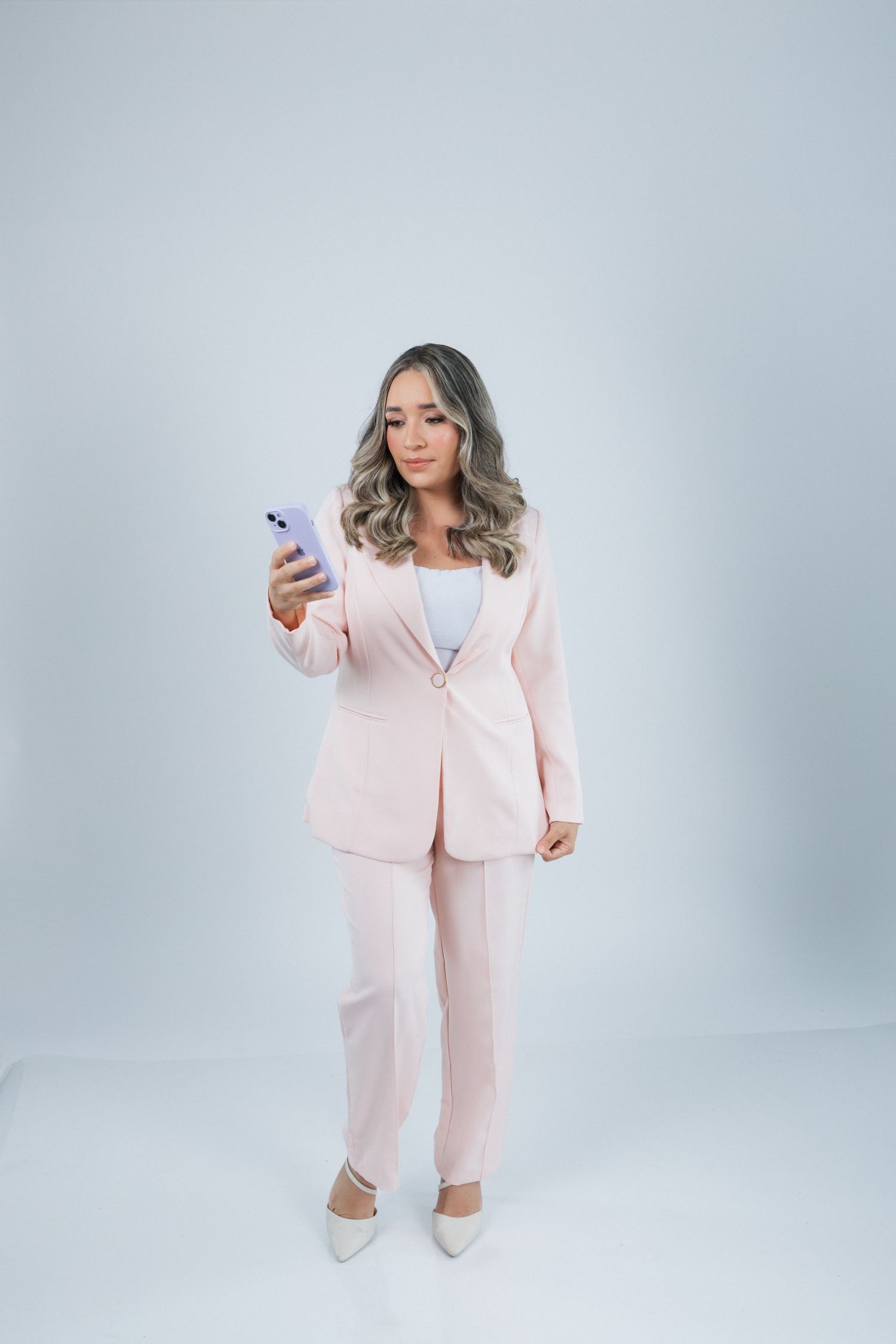 Professional wearing a light pink business suit and heels checks their phone against a white studio background.