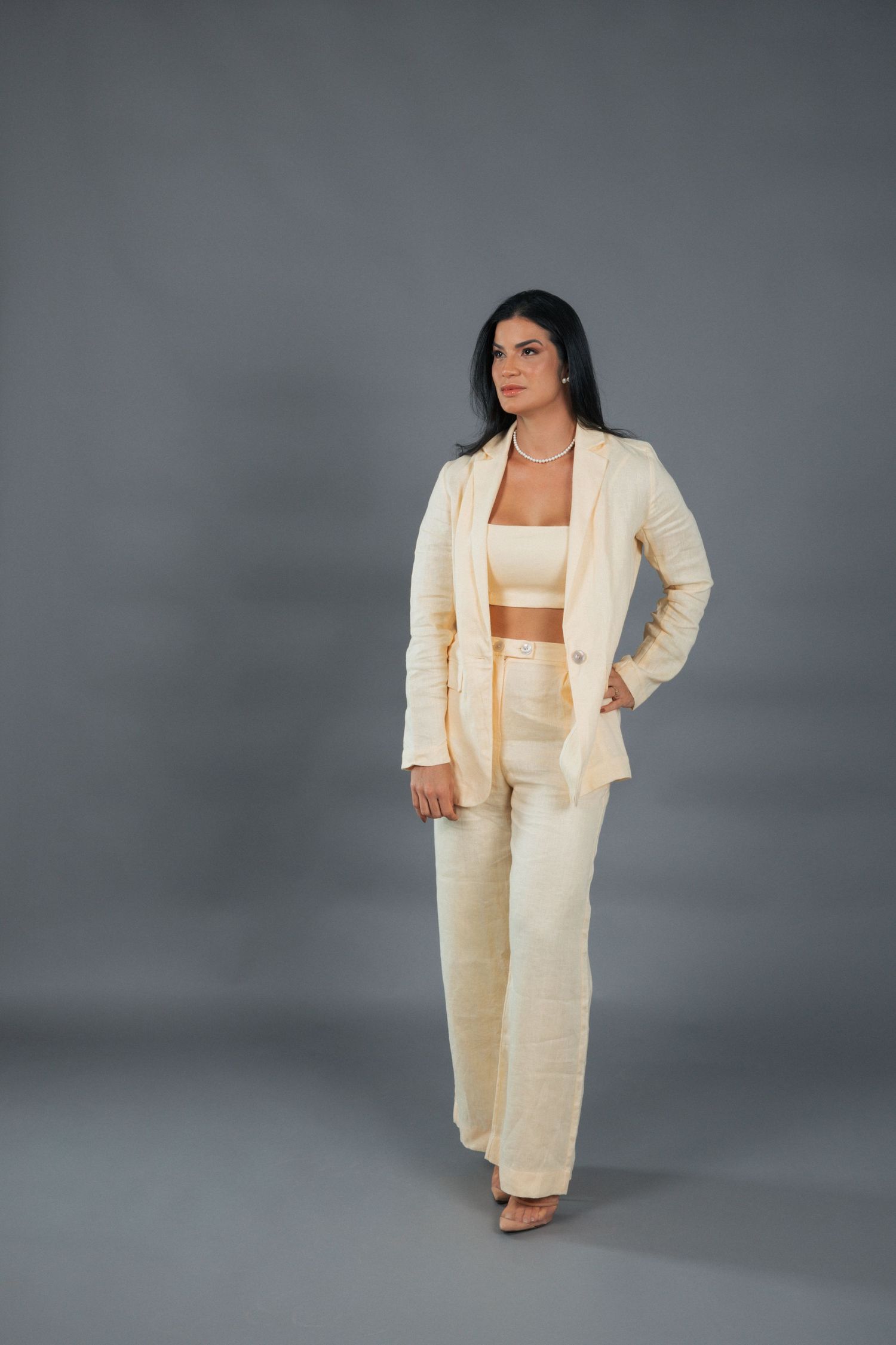 A model wearing a cream-colored three-piece suit with a crop top stands confidently against a gray backdrop.
