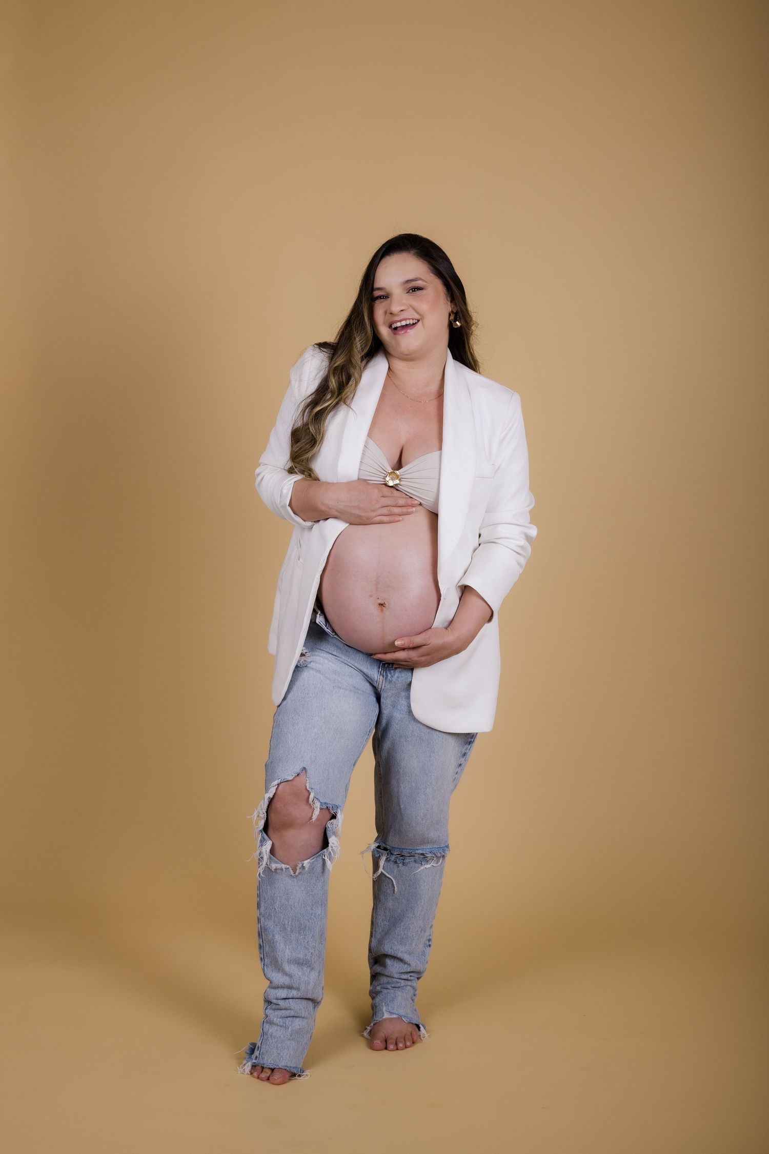 Expectant mother in white blazer and ripped jeans poses against beige backdrop during maternity photoshoot.