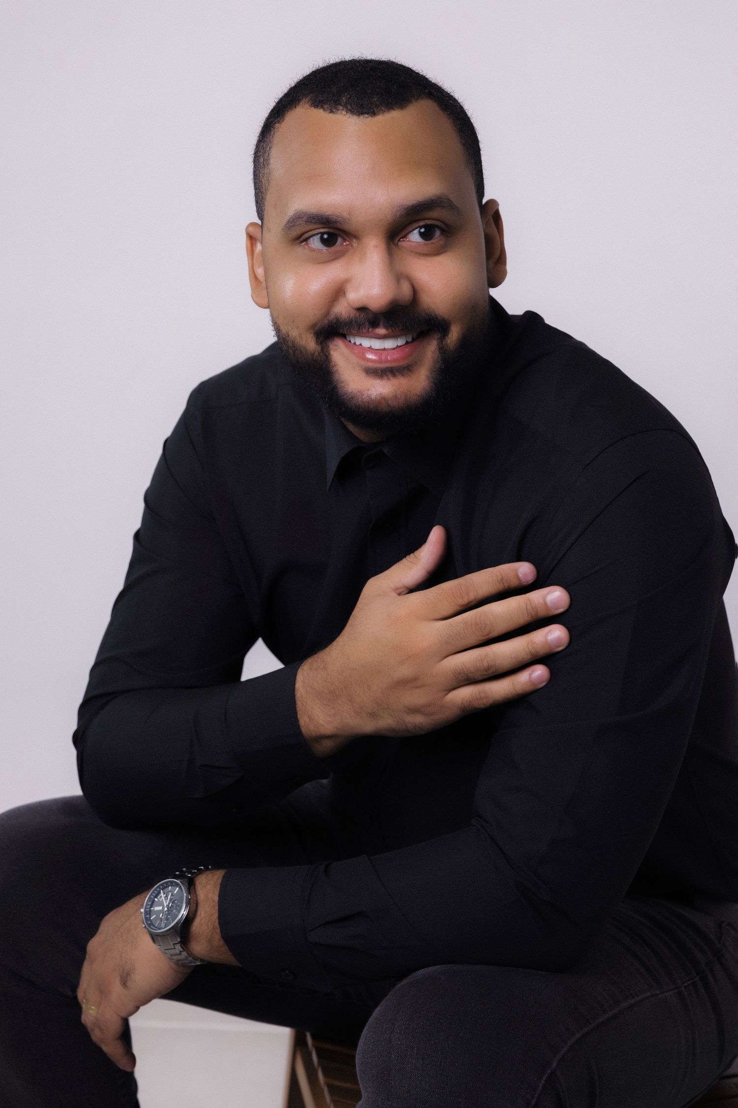 Professional studio portrait of someone in black attire seated against a light gray background with a warm smile.