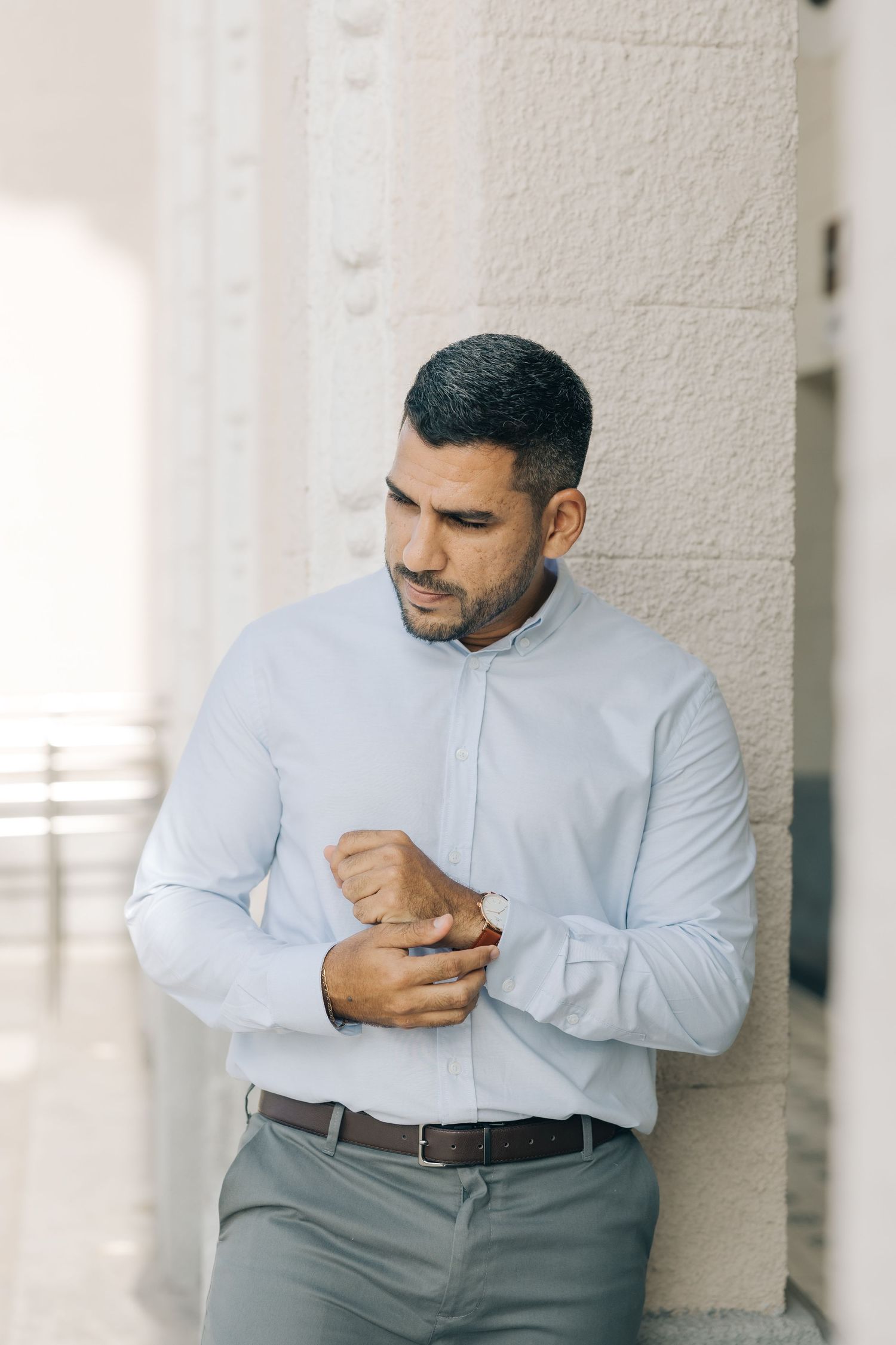 A groom in a light blue dress shirt adjusts his cuffs while standing against a white wall.