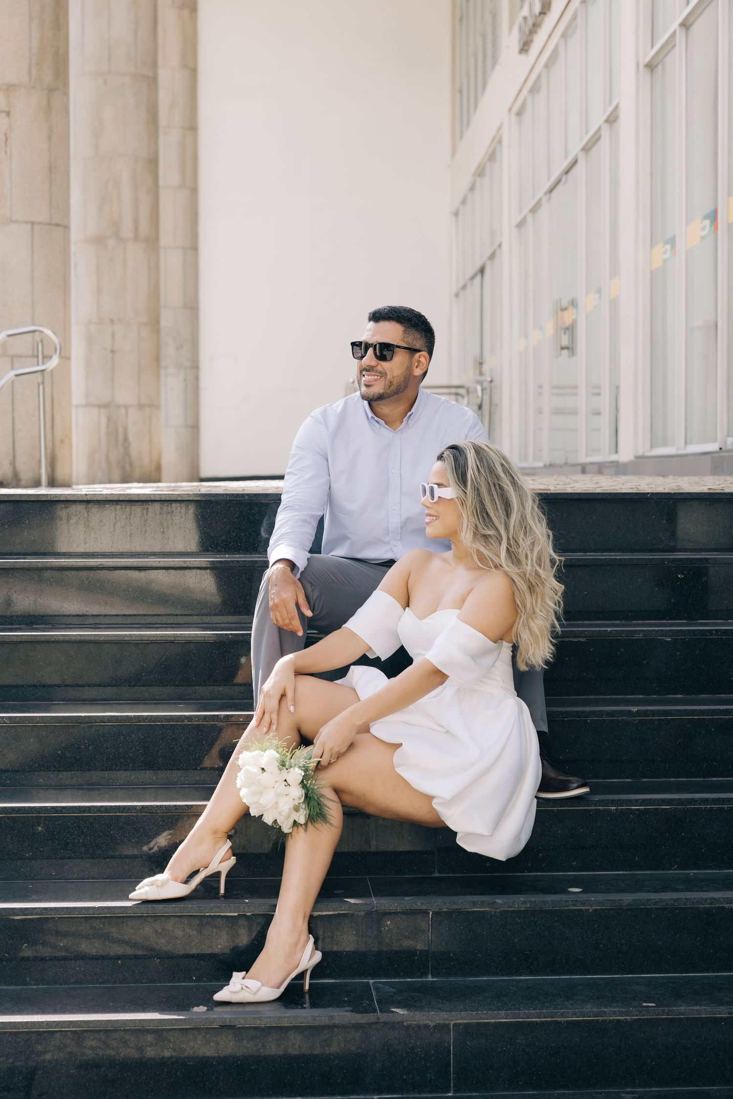A couple sits on black steps outside a building, dressed in elegant casual wear.