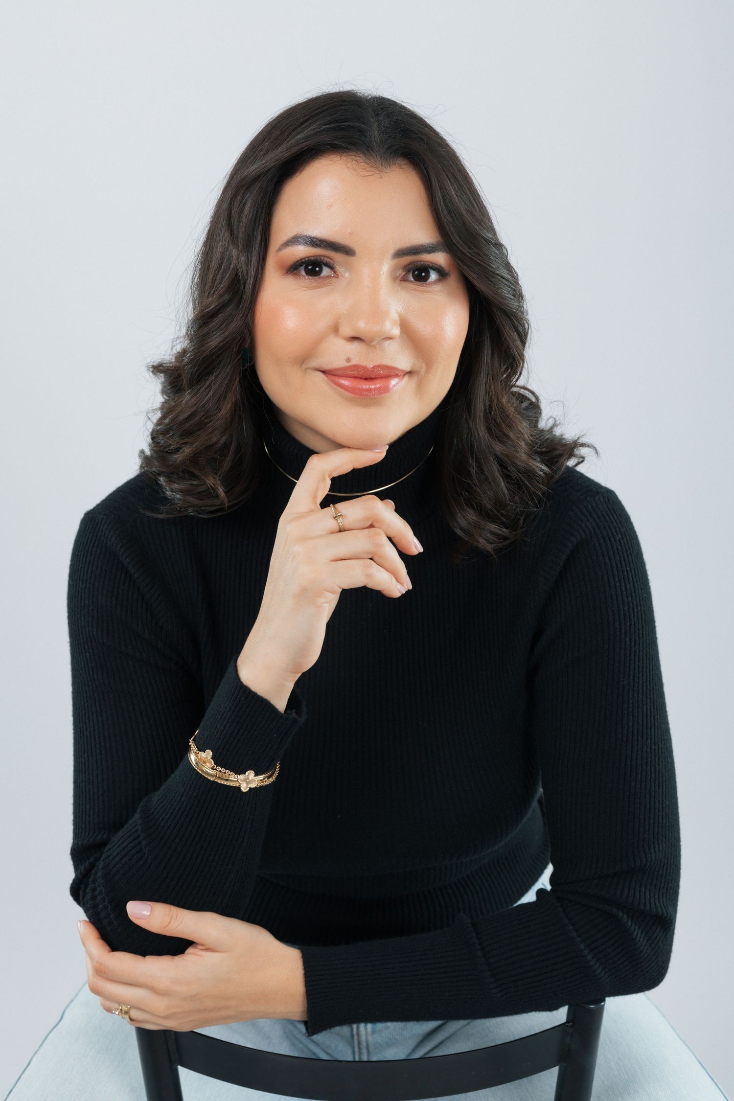 Studio portrait featuring woman in black sweater with contemplative pose and gold bracelet.