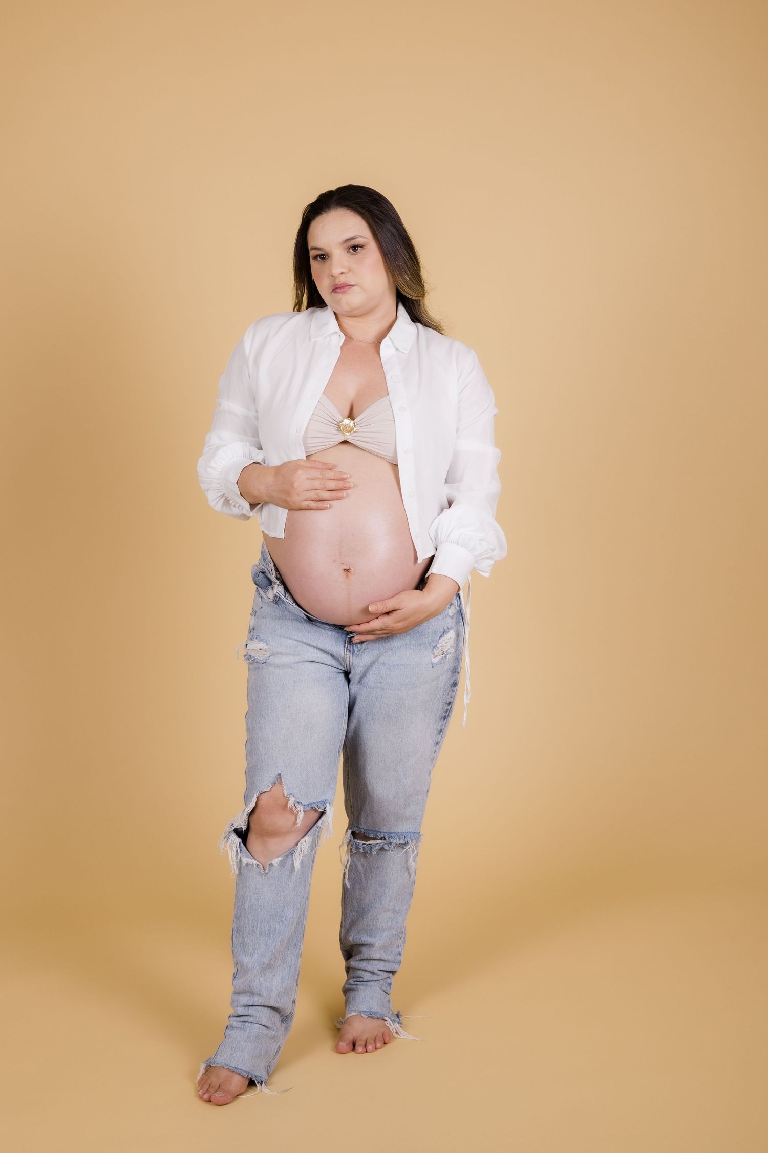 Expectant woman in ripped jeans and white shirt poses against beige backdrop during maternity photoshoot.