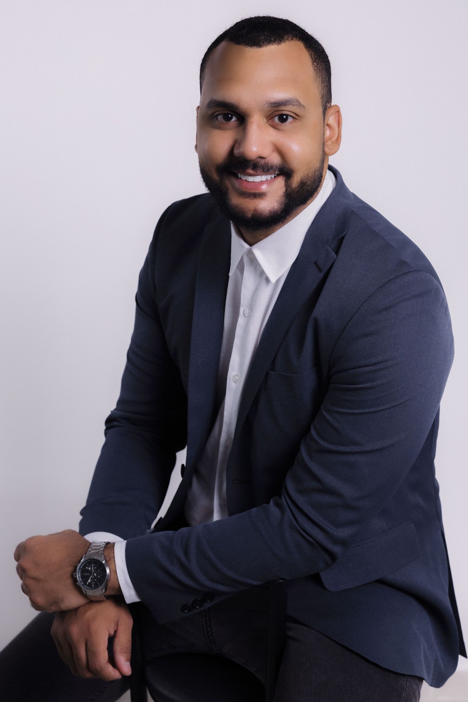 Professional headshot of someone in a navy suit and white shirt seated against a light background with a warm smile.
