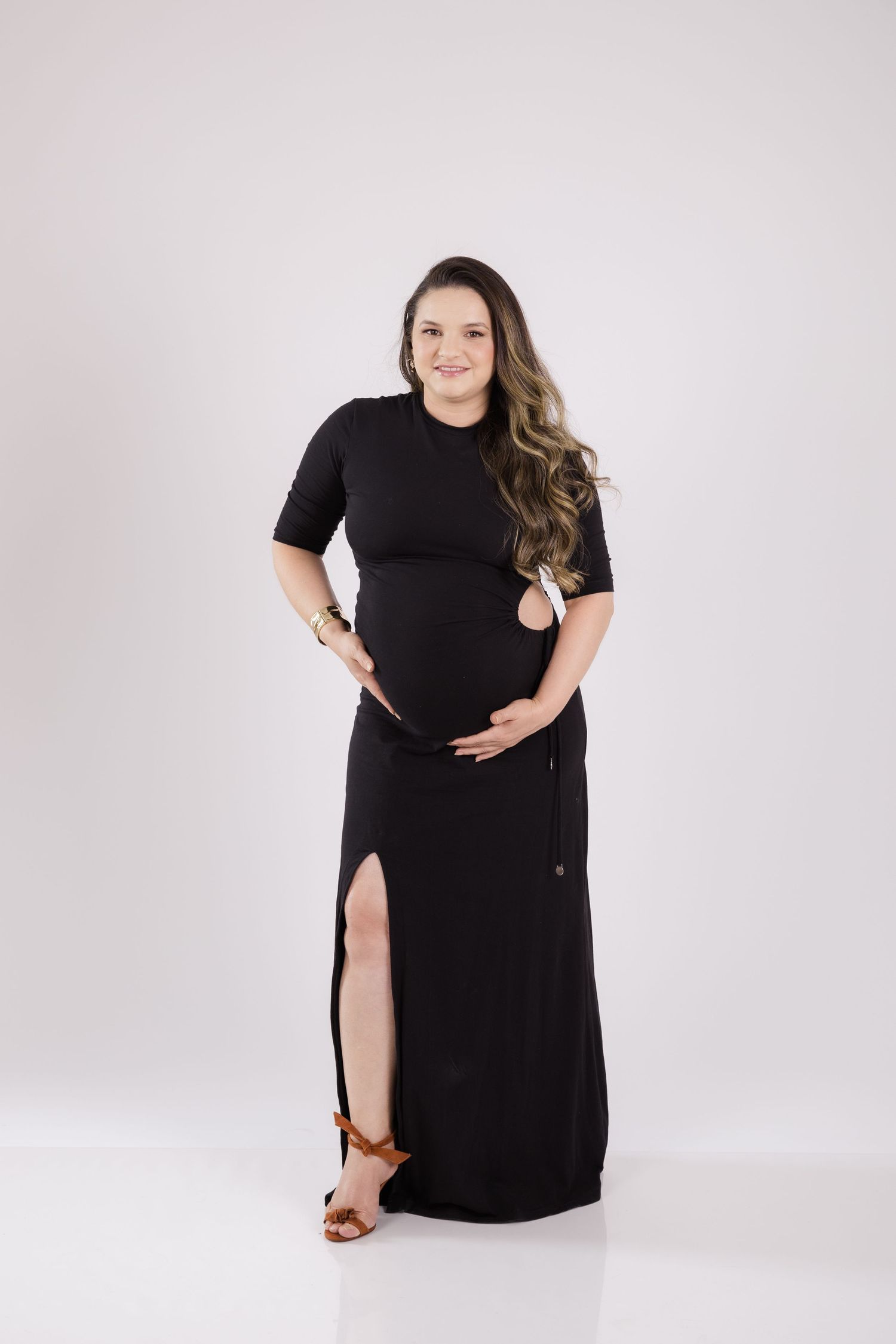 A pregnant woman in a black maxi dress with side slit poses against a light gray backdrop, wearing brown sandals.