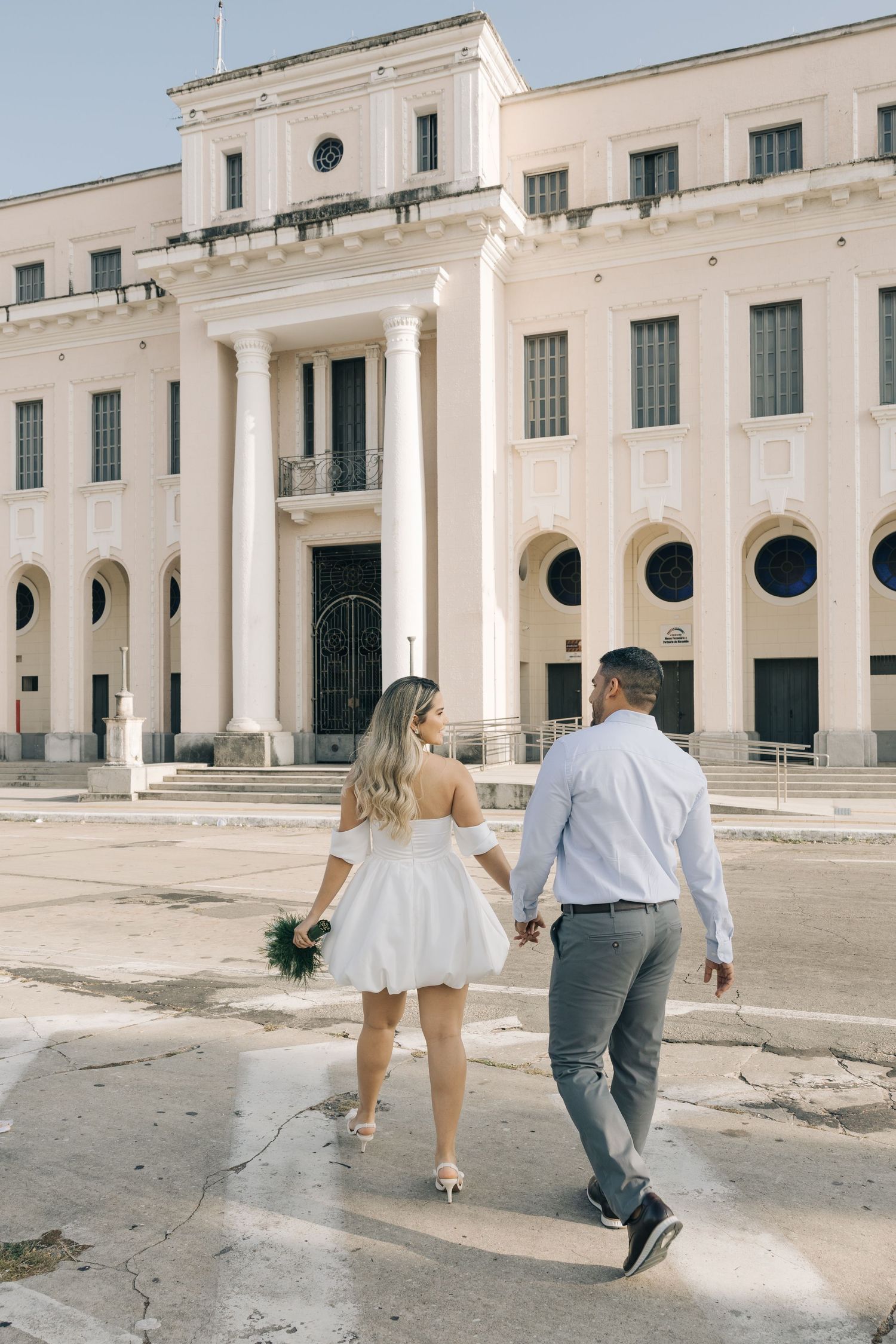 A couple walks hand in hand away from a grand colonial-style building with arched windows and white pillars.