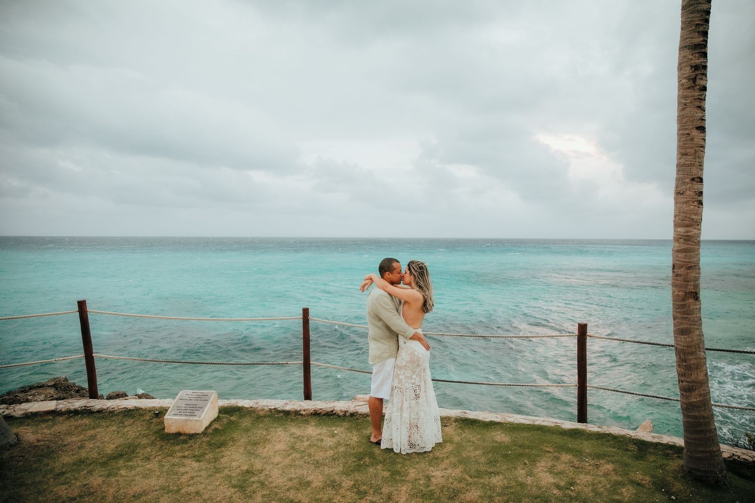 Two people embrace by the ocean's edge on a cloudy day with turquoise waters and a palm tree in view.