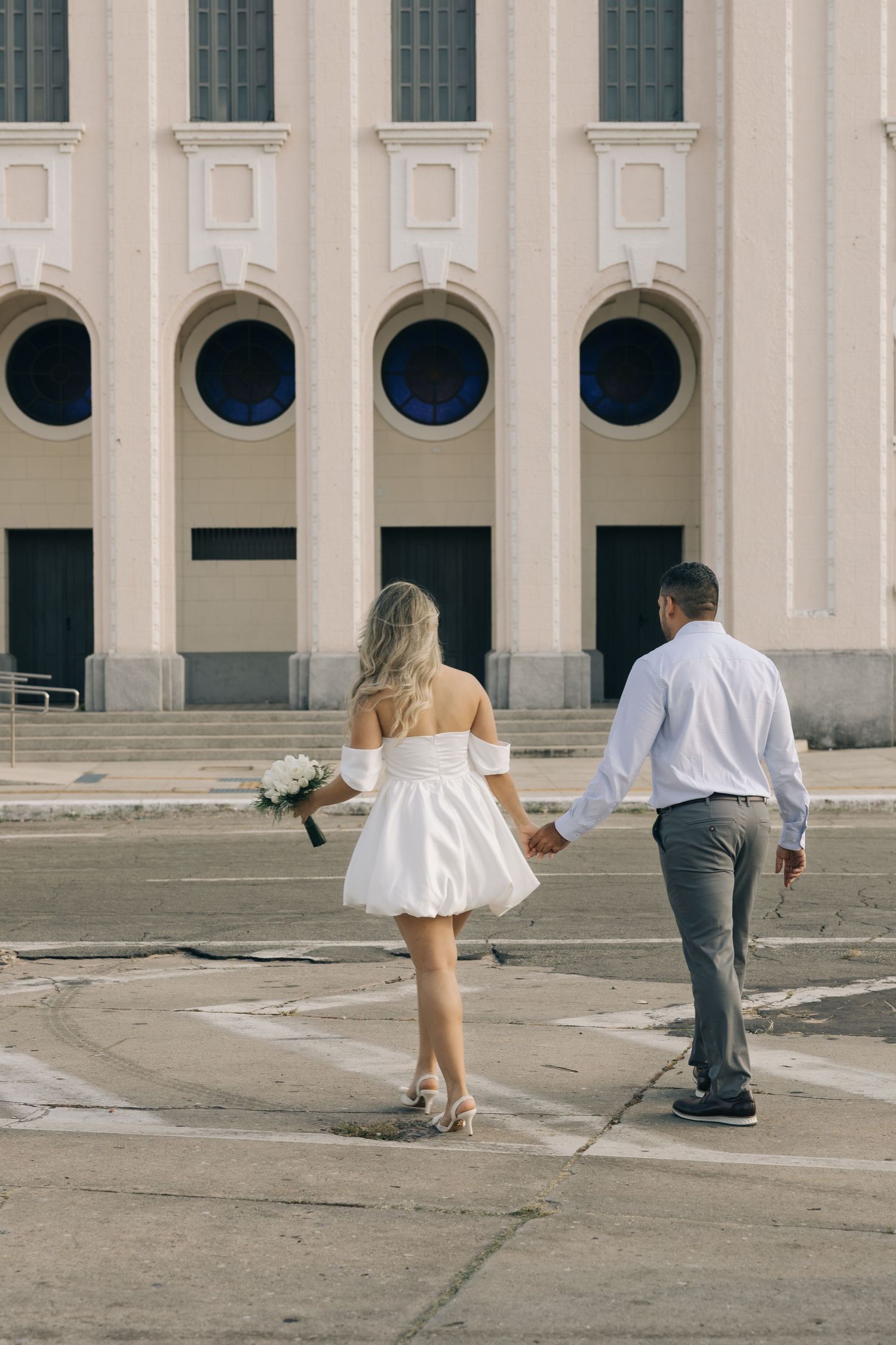 A couple walks together holding hands near a building with arched windows and circular portals.