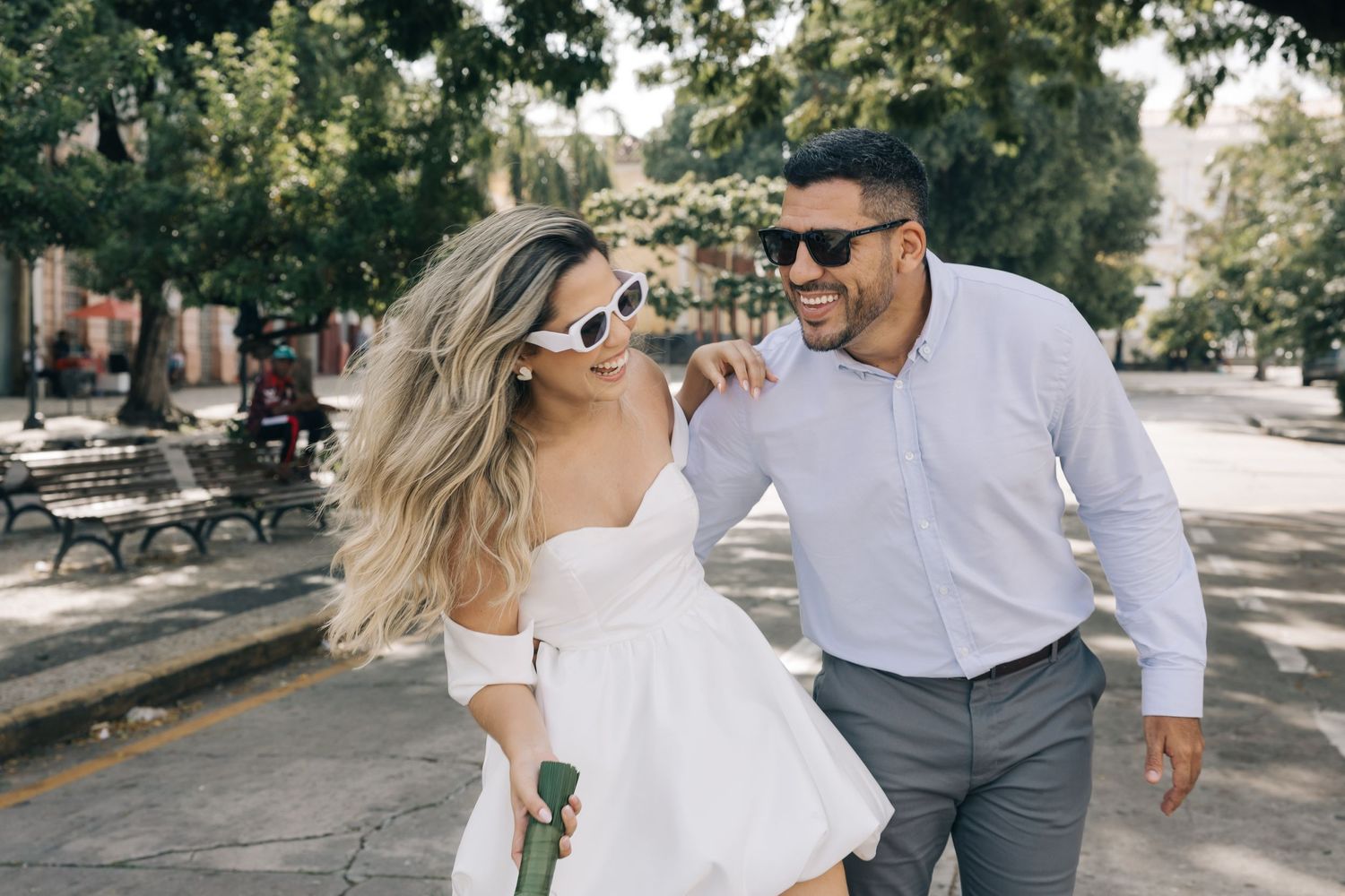 A couple in white clothing share a joyful moment while walking down a tree-lined street.