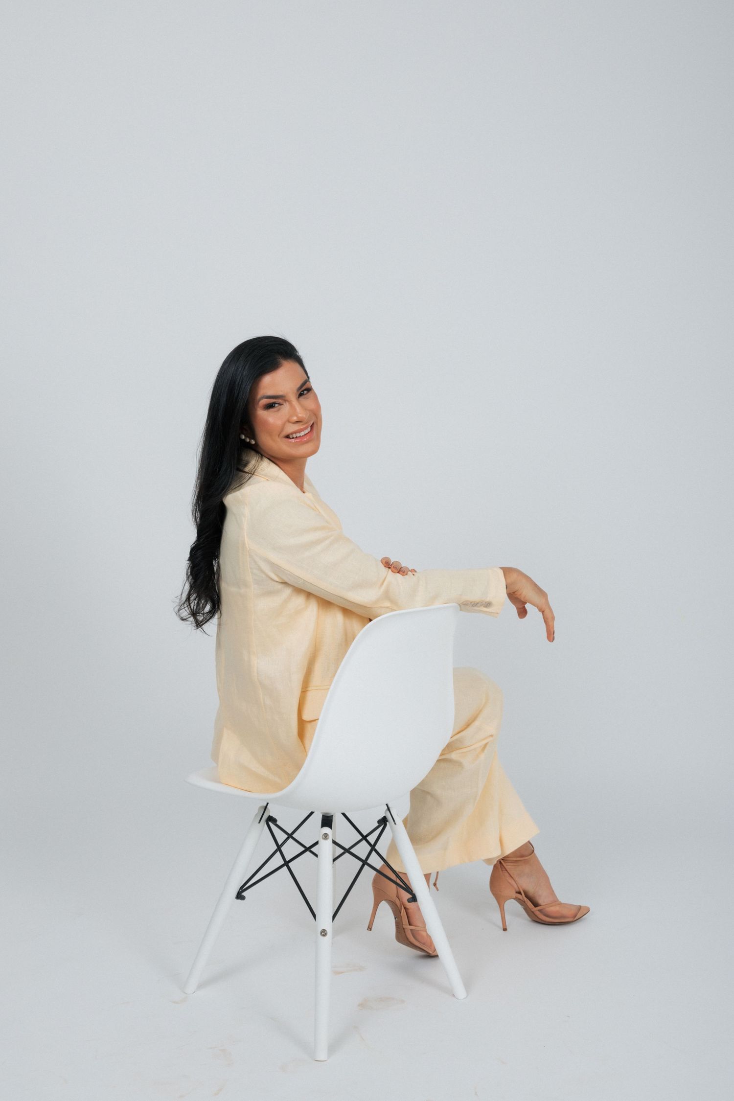 Woman in beige pantsuit sitting on white modern chair in studio setting.