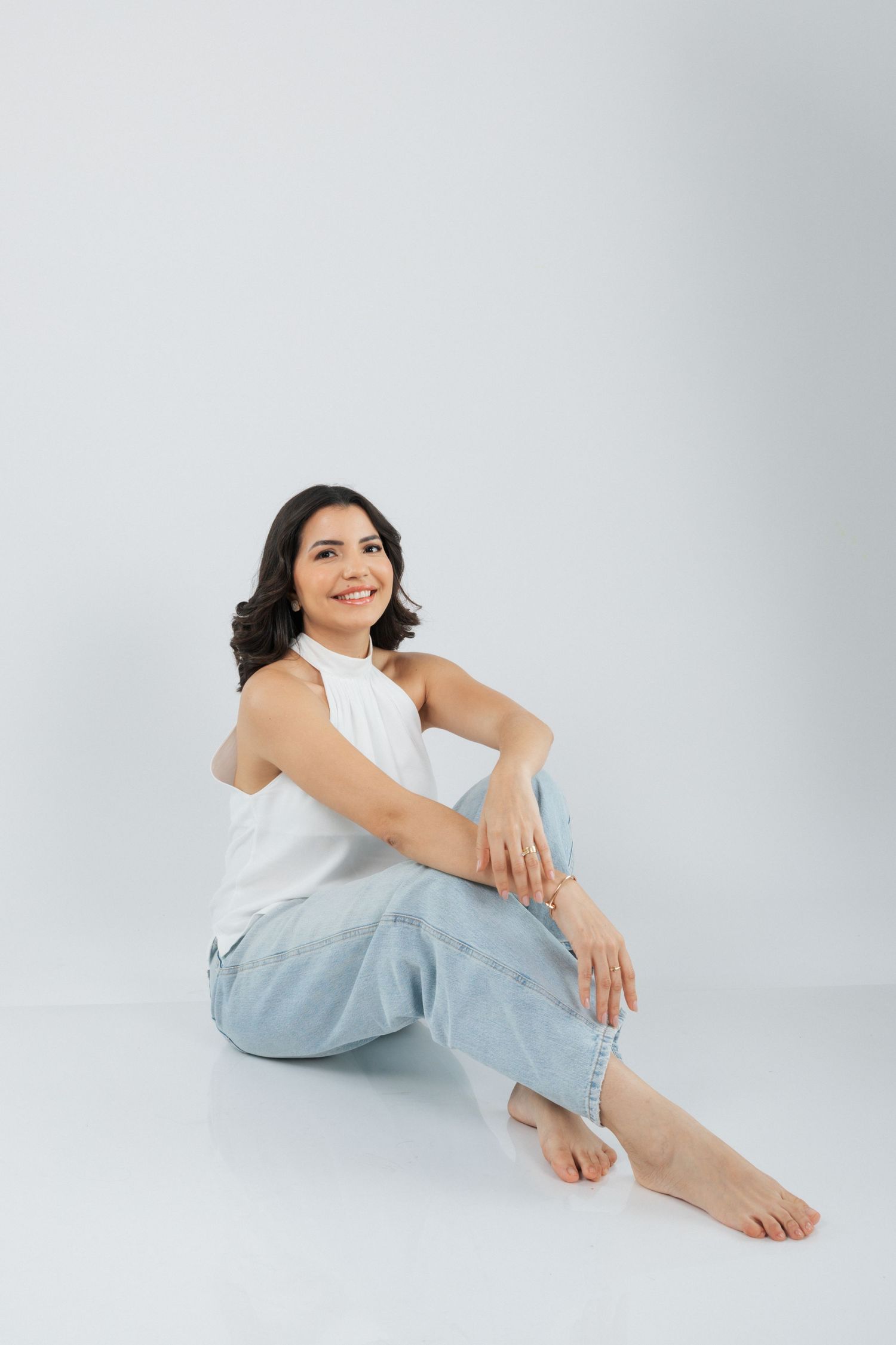 Person in white sleeveless top and light jeans sits casually on white background with relaxed barefoot pose.