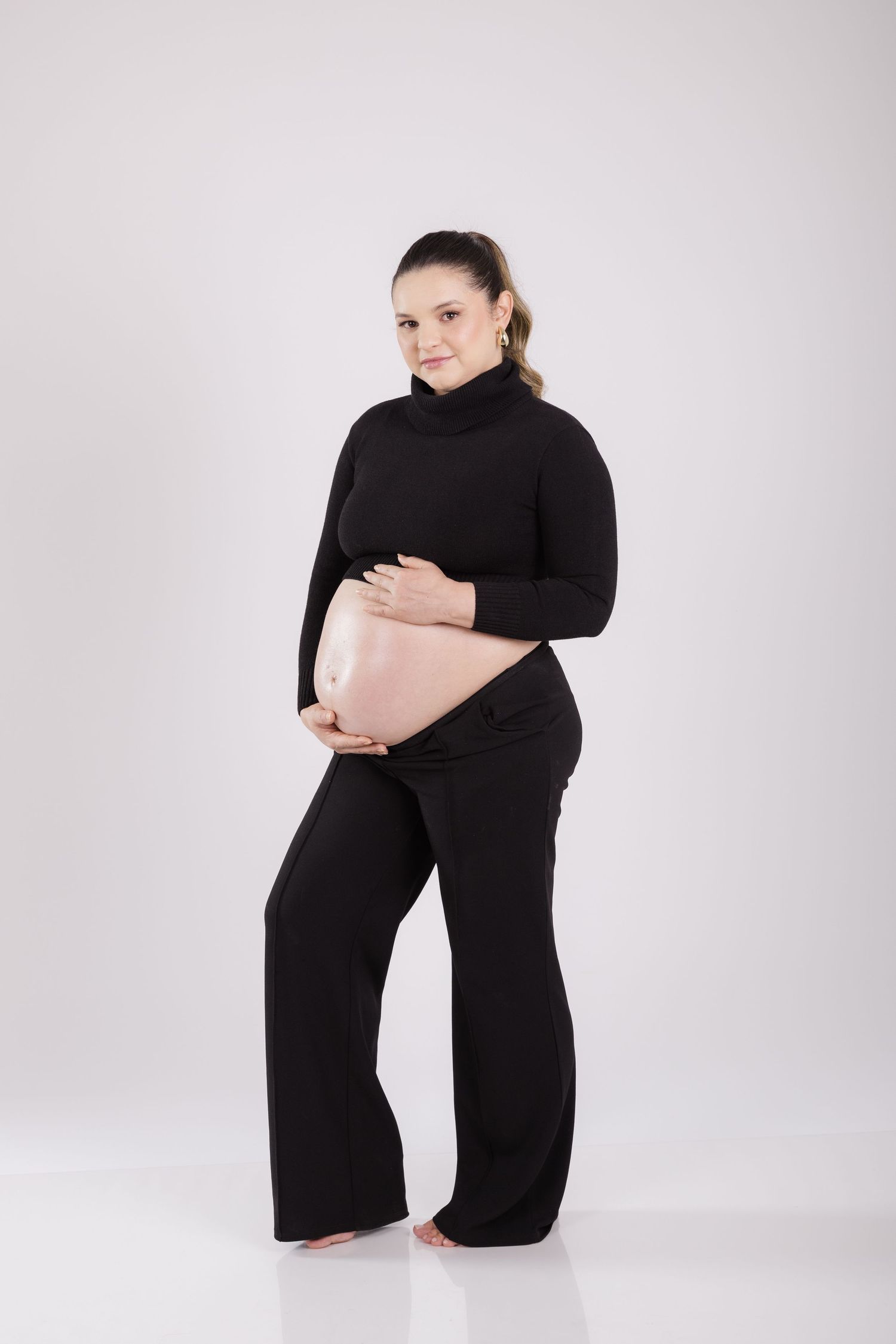 Person in a black turtleneck and wide-leg pants poses against a neutral background, leaning on a pink pillow.