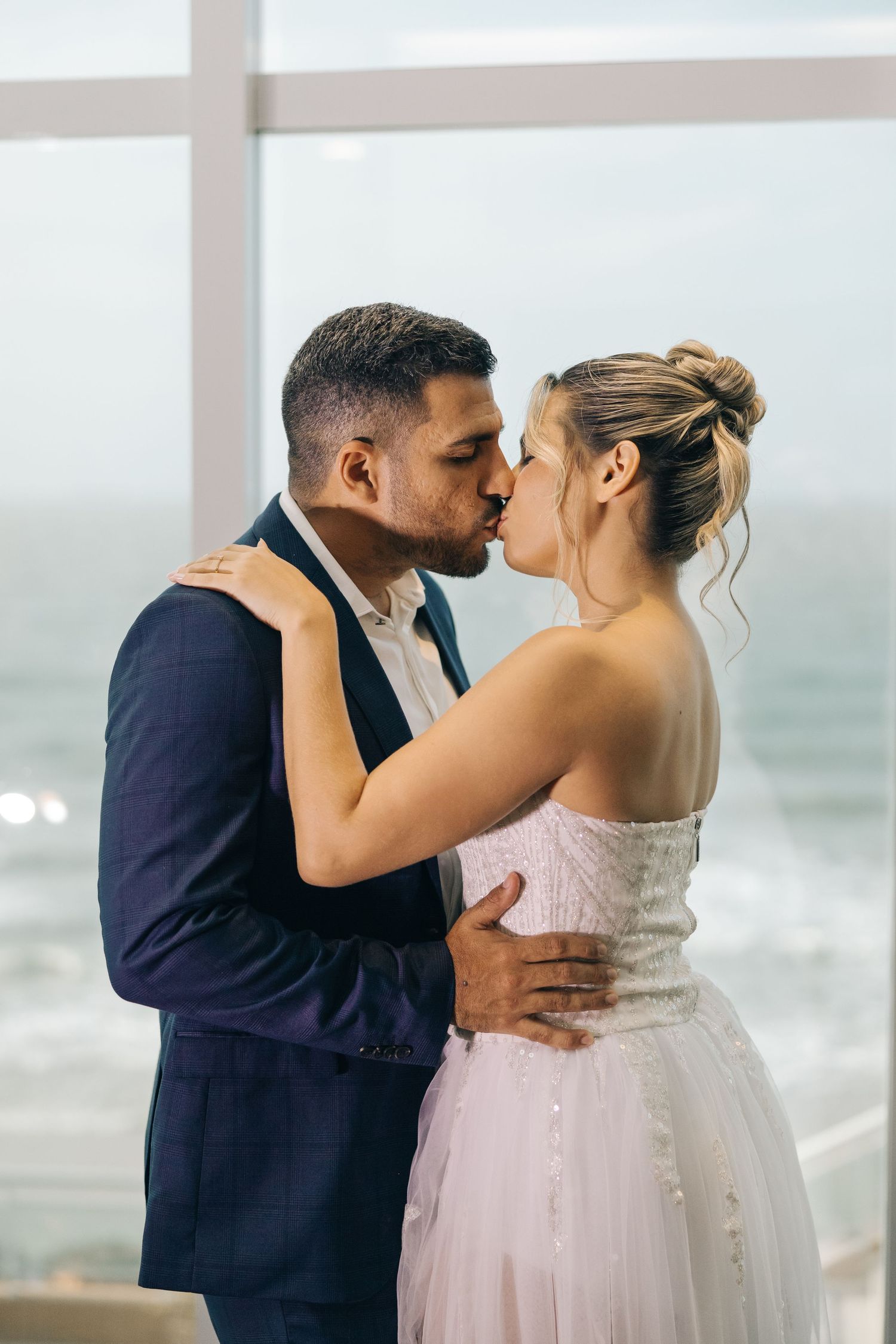 A romantic wedding couple shares a kiss by the ocean, with the bride in a blush gown and groom in navy suit.