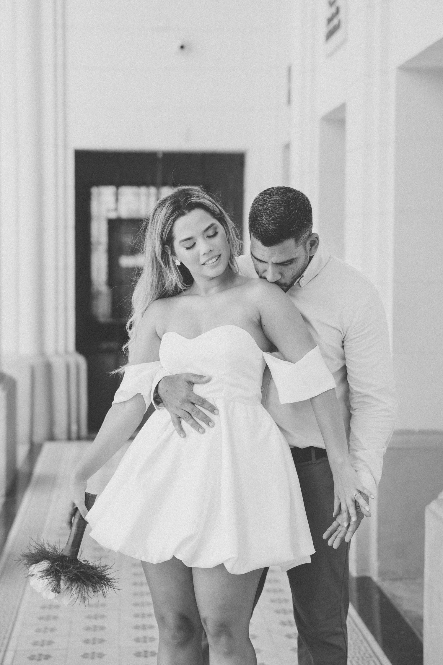 A romantic black and white photo of a couple embracing in a white hallway, with the woman wearing a short white dress.