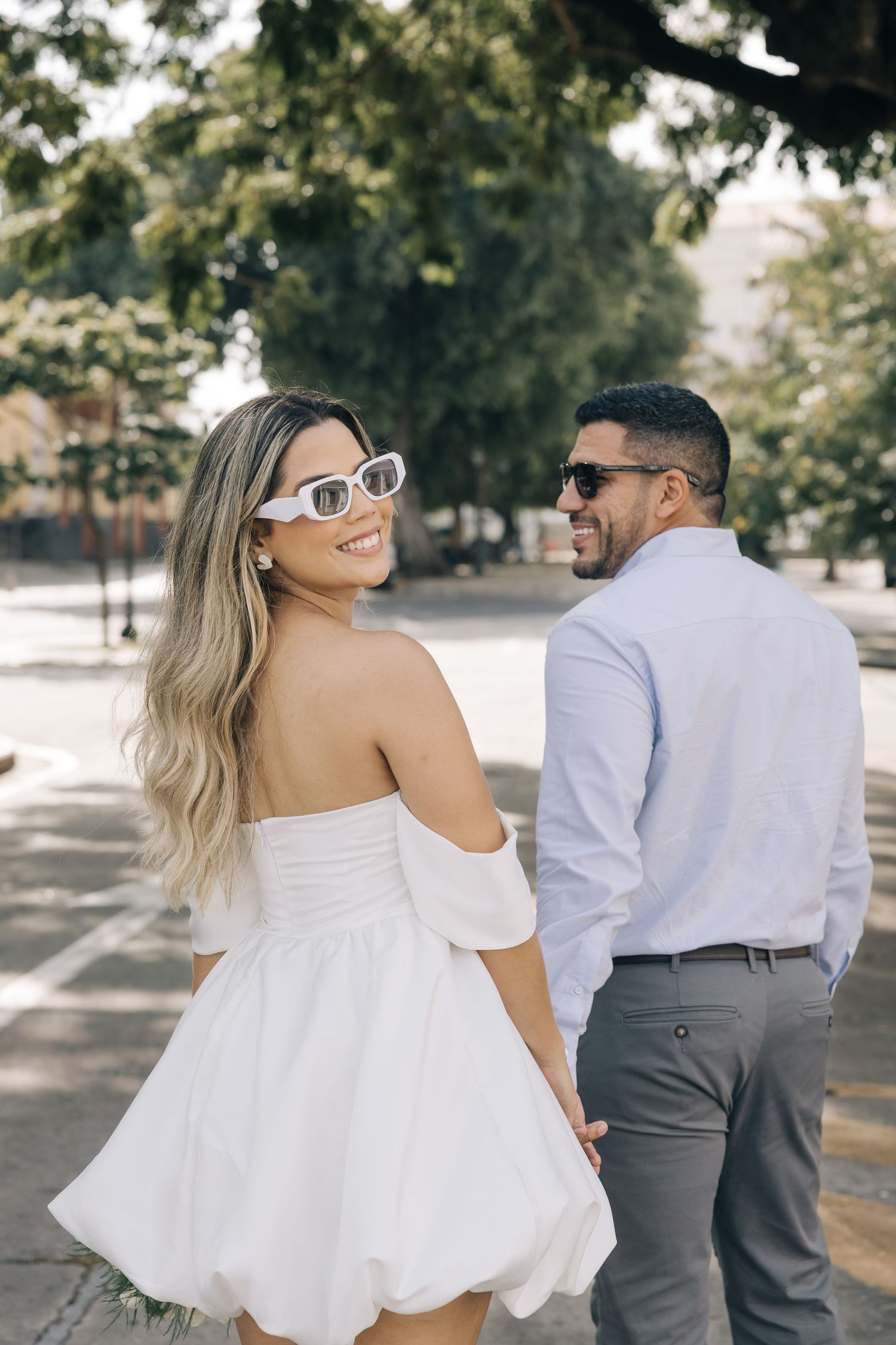 A couple in coordinated white and grey outfits sharing a playful moment under trees.
