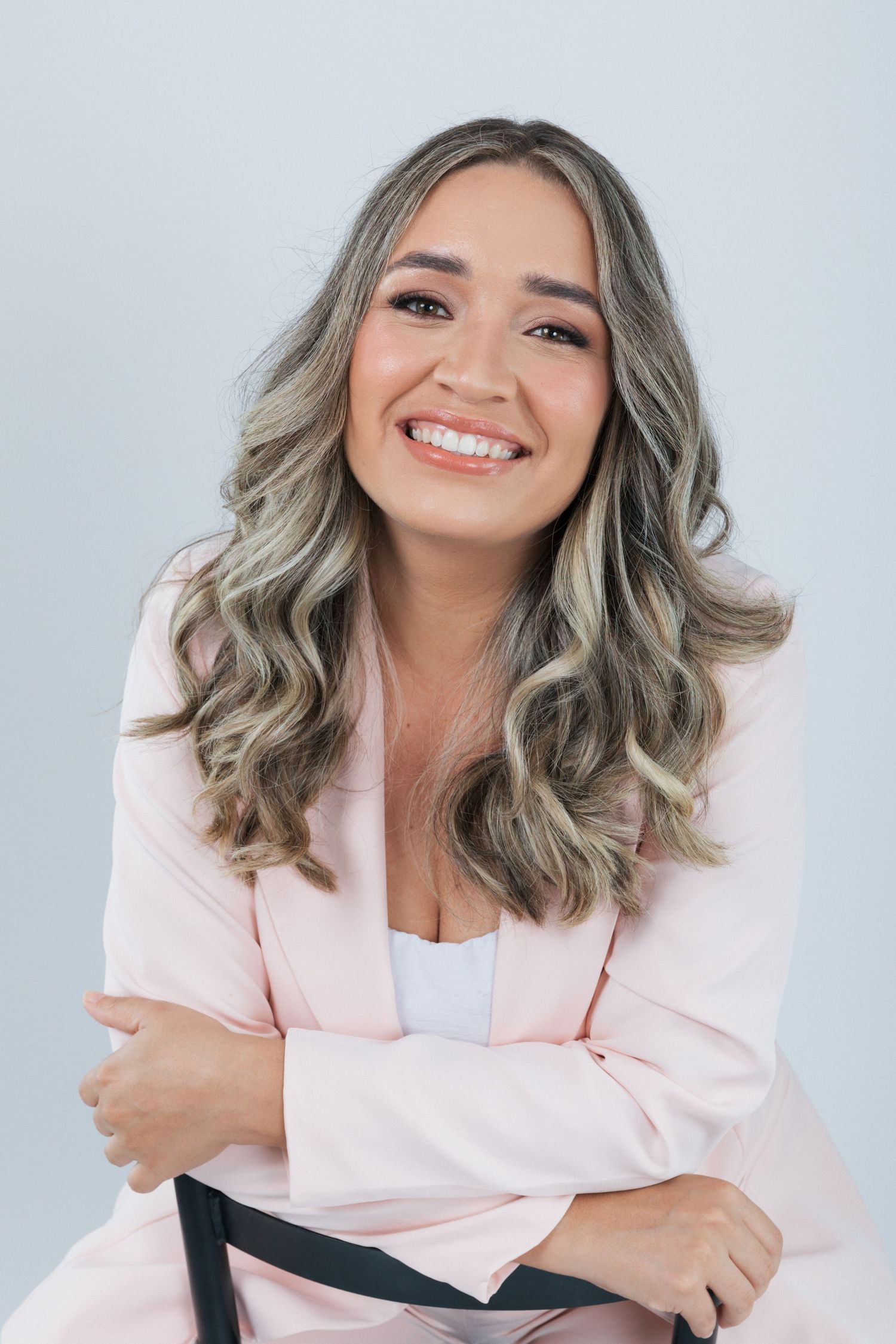 Professional portrait of a person wearing a pink blazer and white top with styled grey wavy hair against light background.