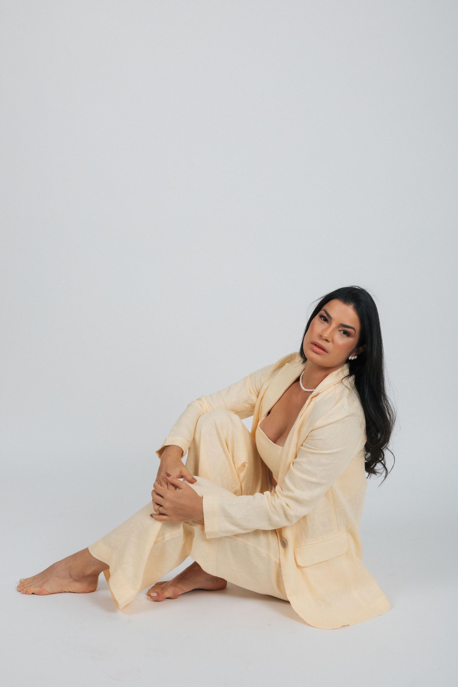 Model poses sitting on white backdrop wearing light cream colored blazer and pants with pearl necklace.