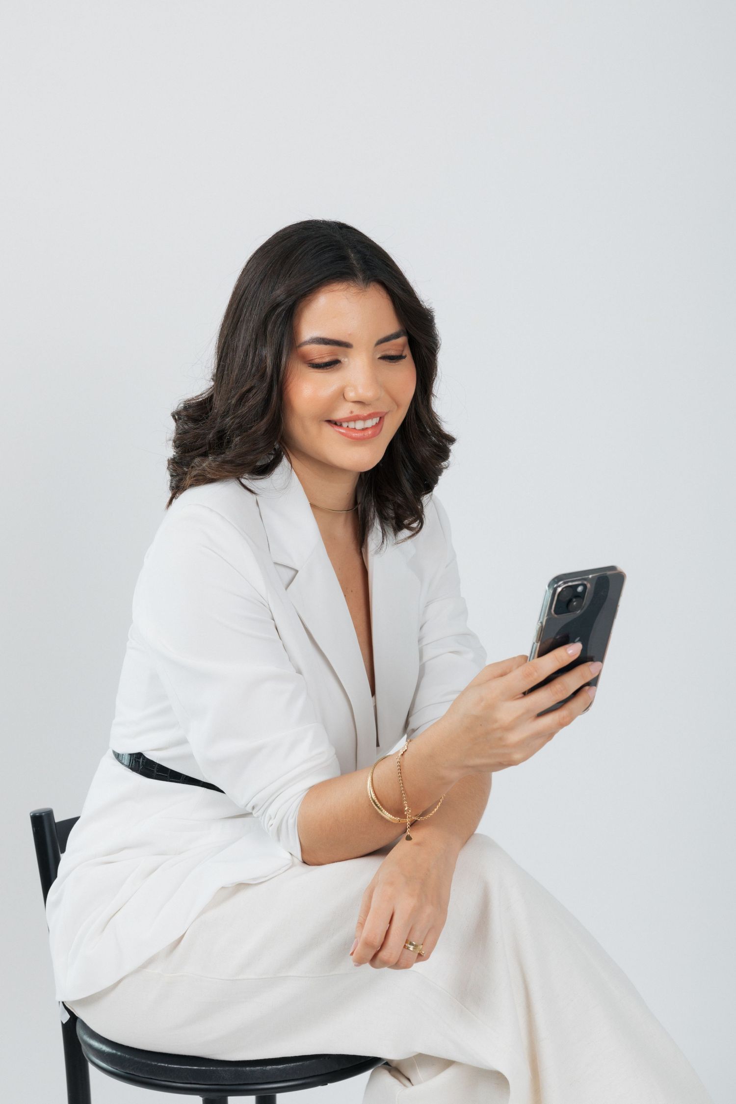Professional wearing a white blazer smiles while looking at smartphone screen on grey background.