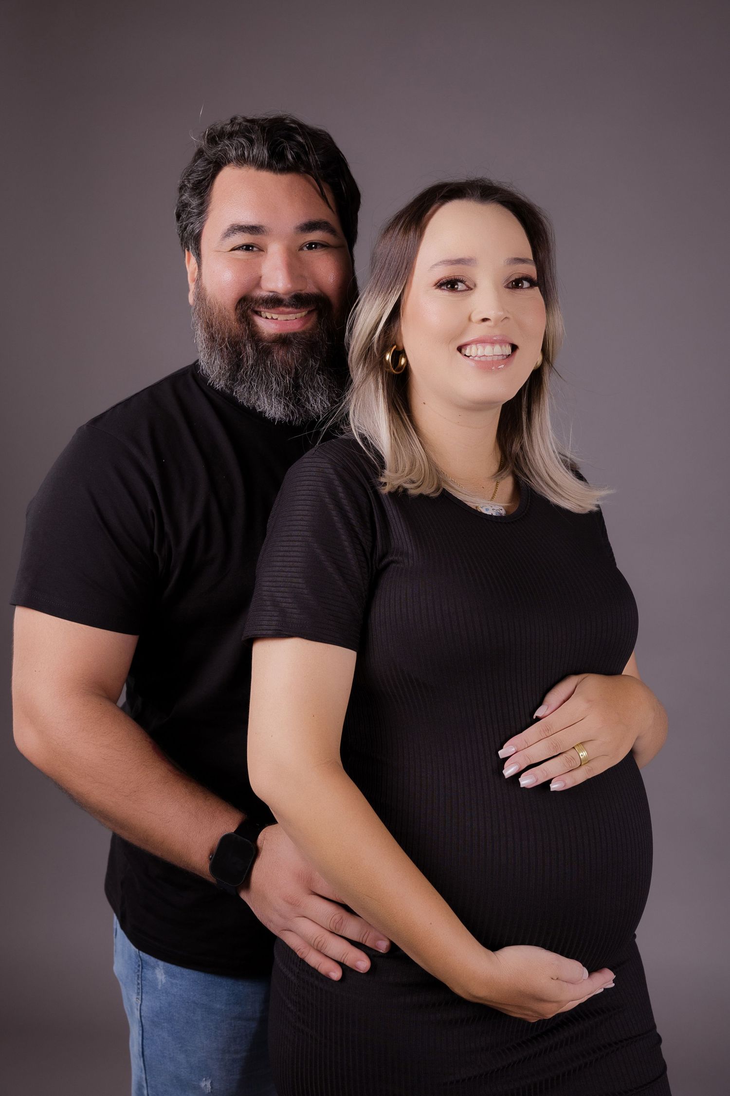 An expectant couple in matching black shirts pose together against a dark grey studio background.