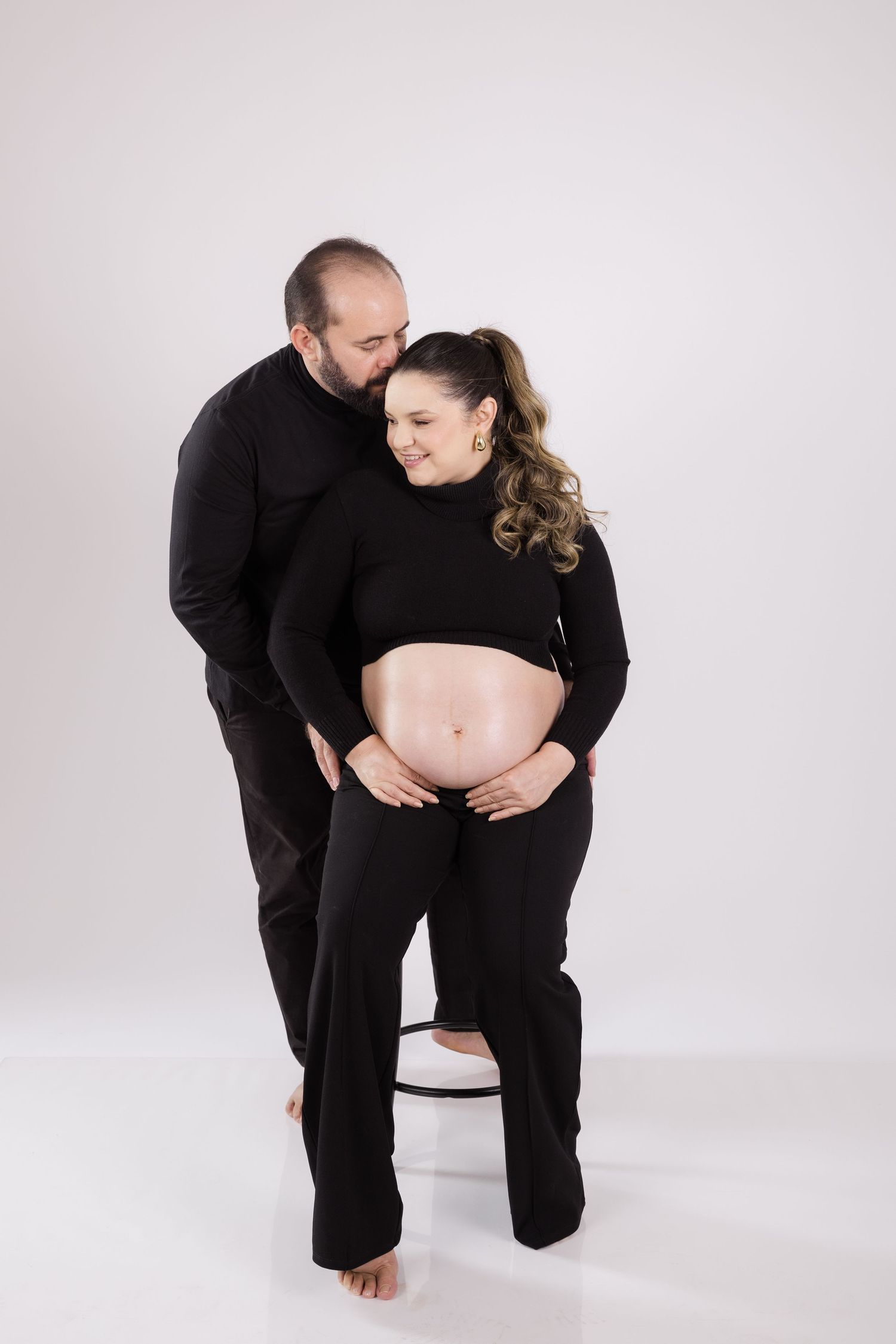 Tender pregnancy portrait shows a couple dressed in black as they share a loving moment against light background.