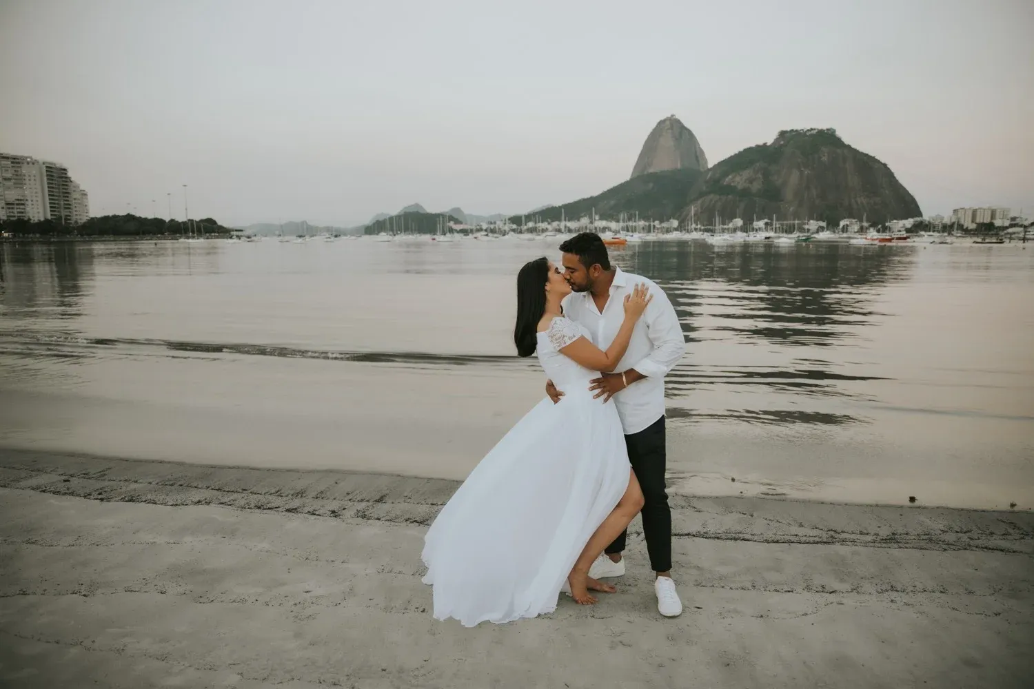 A couple in wedding attire share a romantic moment on the beach with Rio de Janeiro's Sugarloaf Mountain in the background.