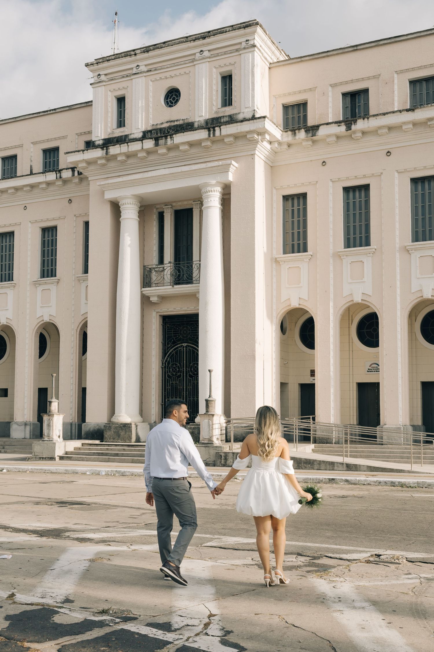 A couple holding hands walking in front of a grand white neoclassical building with columns and circular windows.