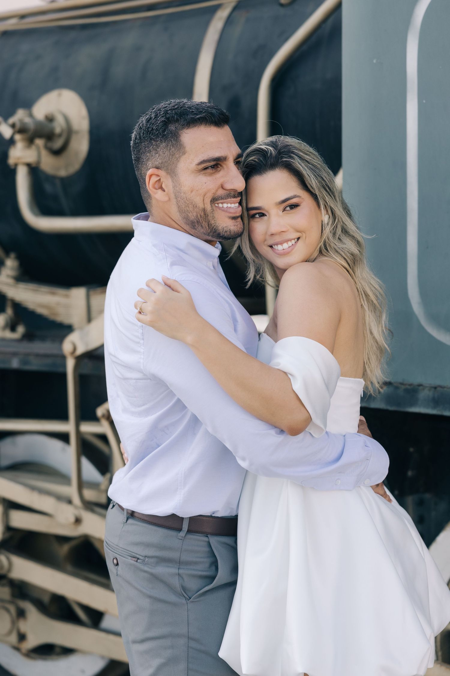A couple in elegant attire embrace and smile beside a vintage train car during an outdoor photo session.
