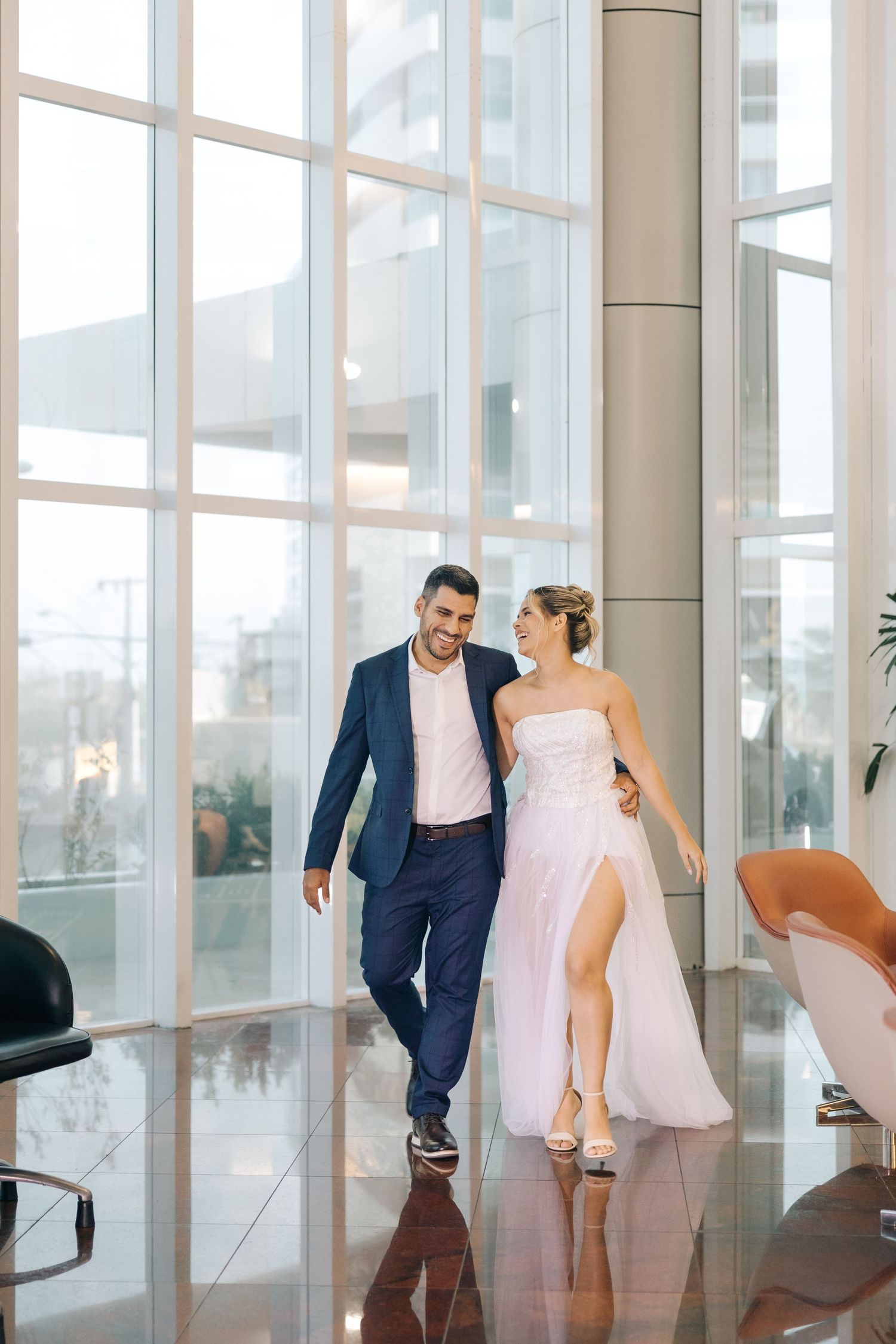 A couple walks together in formal wear through a modern building with floor-to-ceiling windows.